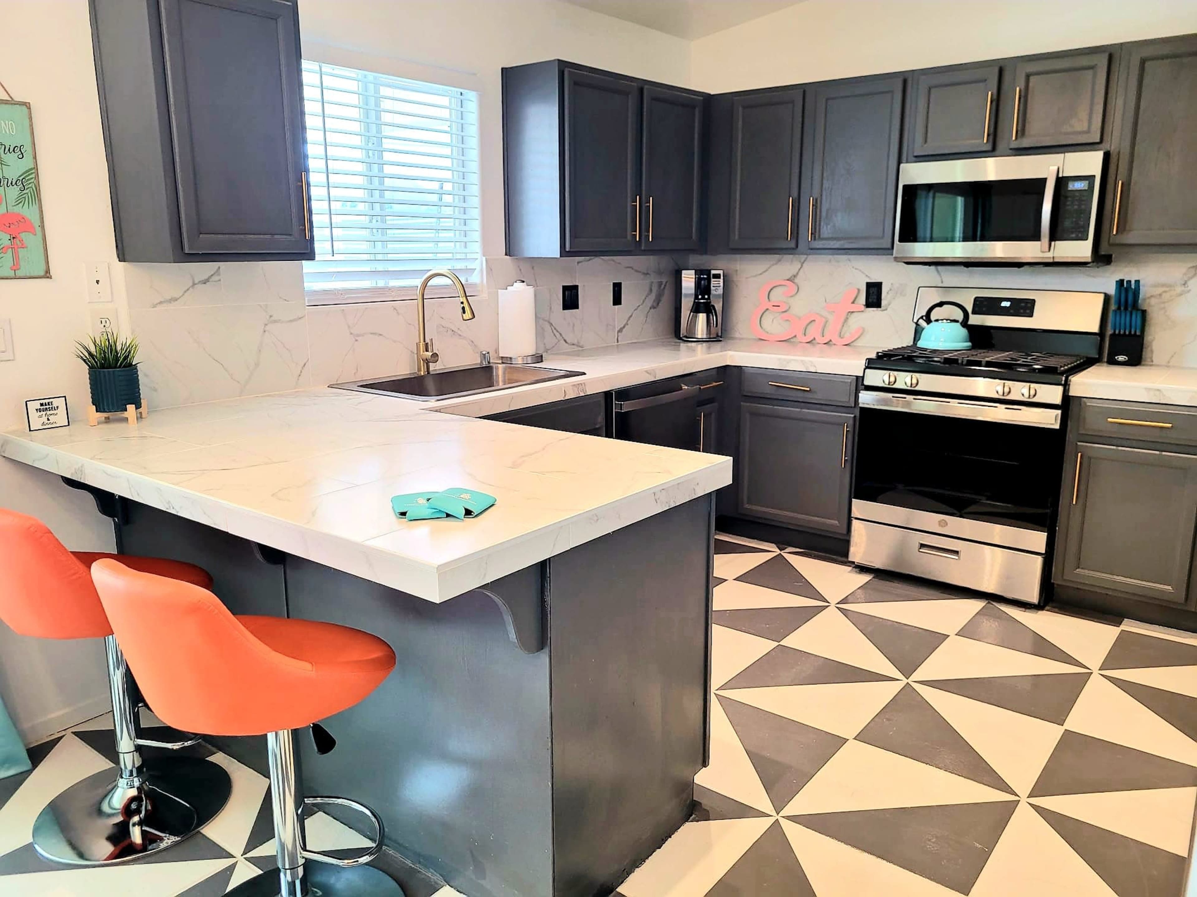 A modern kitchen features gray cabinetry, stainless steel appliances, a marble countertop, and orange bar stools at a tiled breakfast bar.