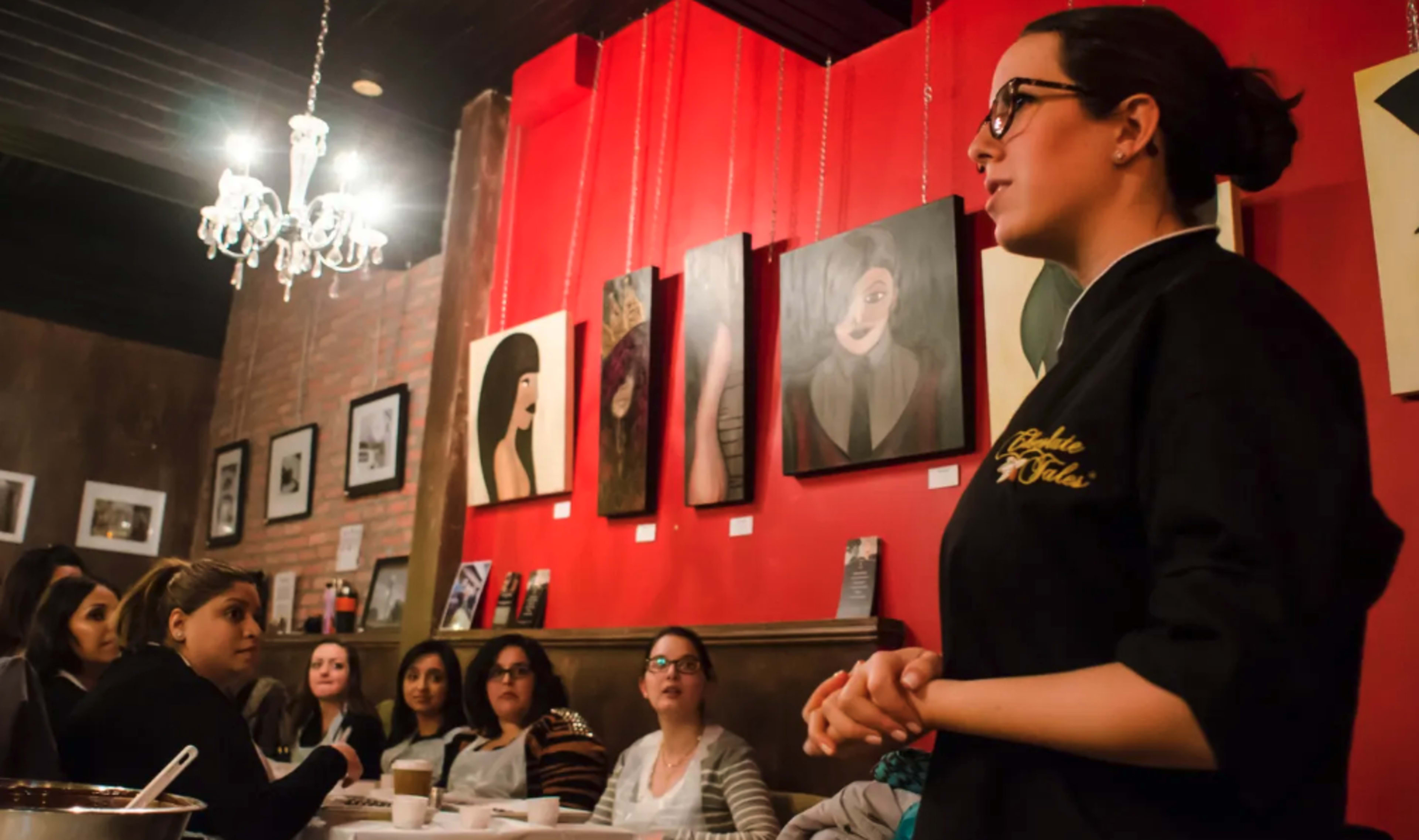 A woman in a black jacket speaks to a seated audience in a café adorned with various paintings on the walls.
