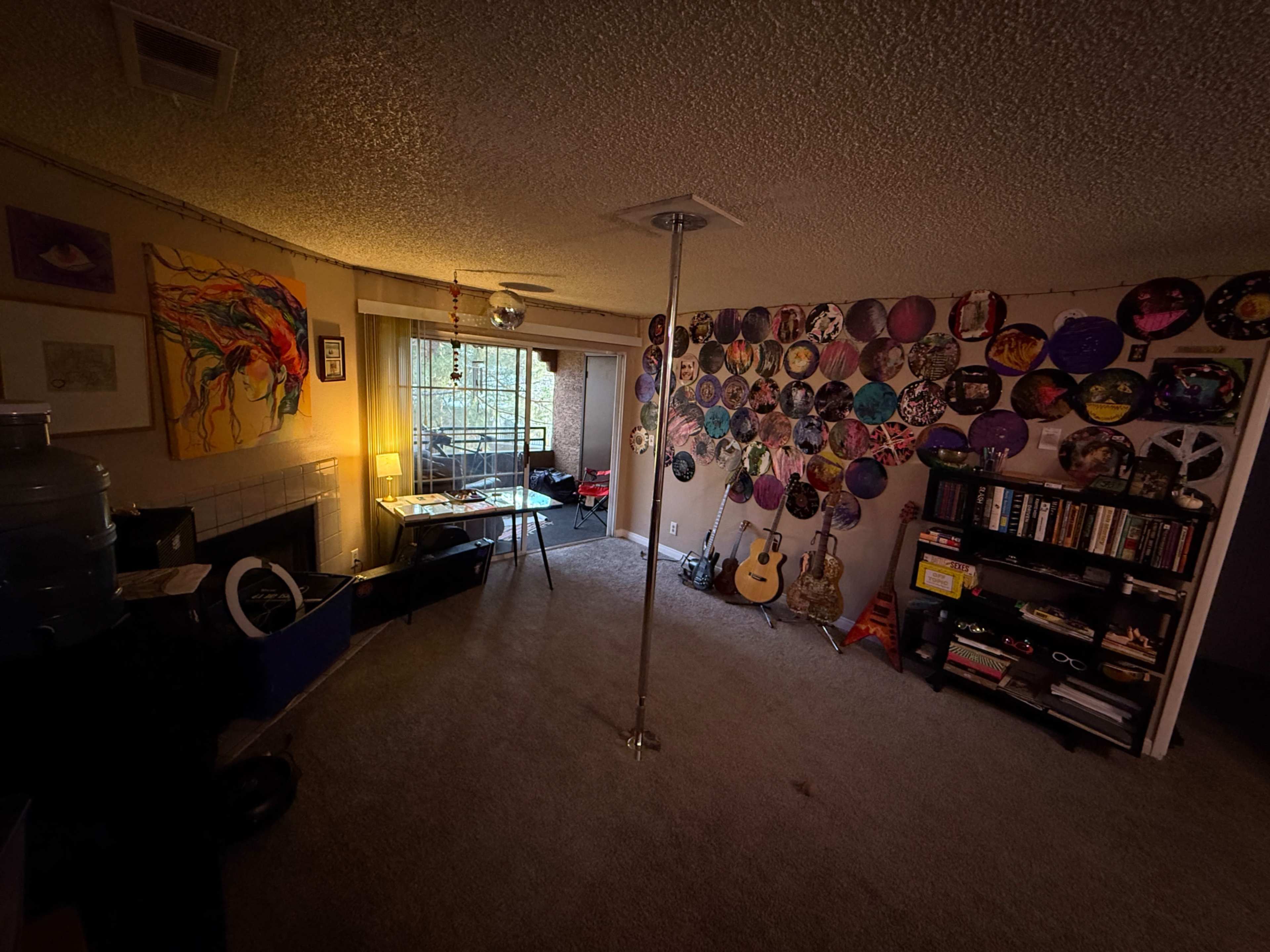 The image depicts a cozy living space with a pole in the center, a table and chairs near a window, and walls adorned with colorful vinyl records and artwork.