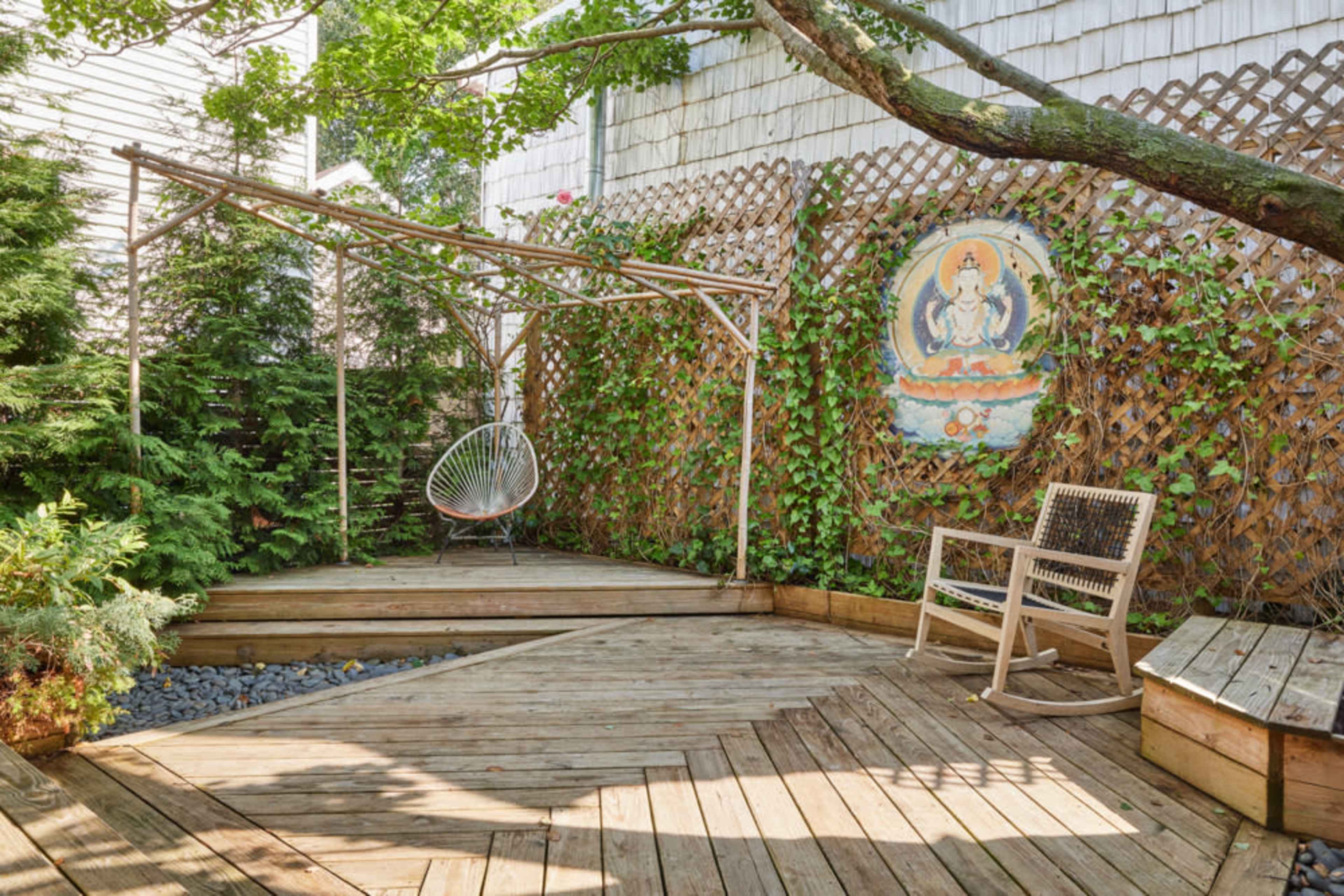 A wooden deck surrounded by greenery, featuring a hanging chair and a decorative wall with an ornate painting.