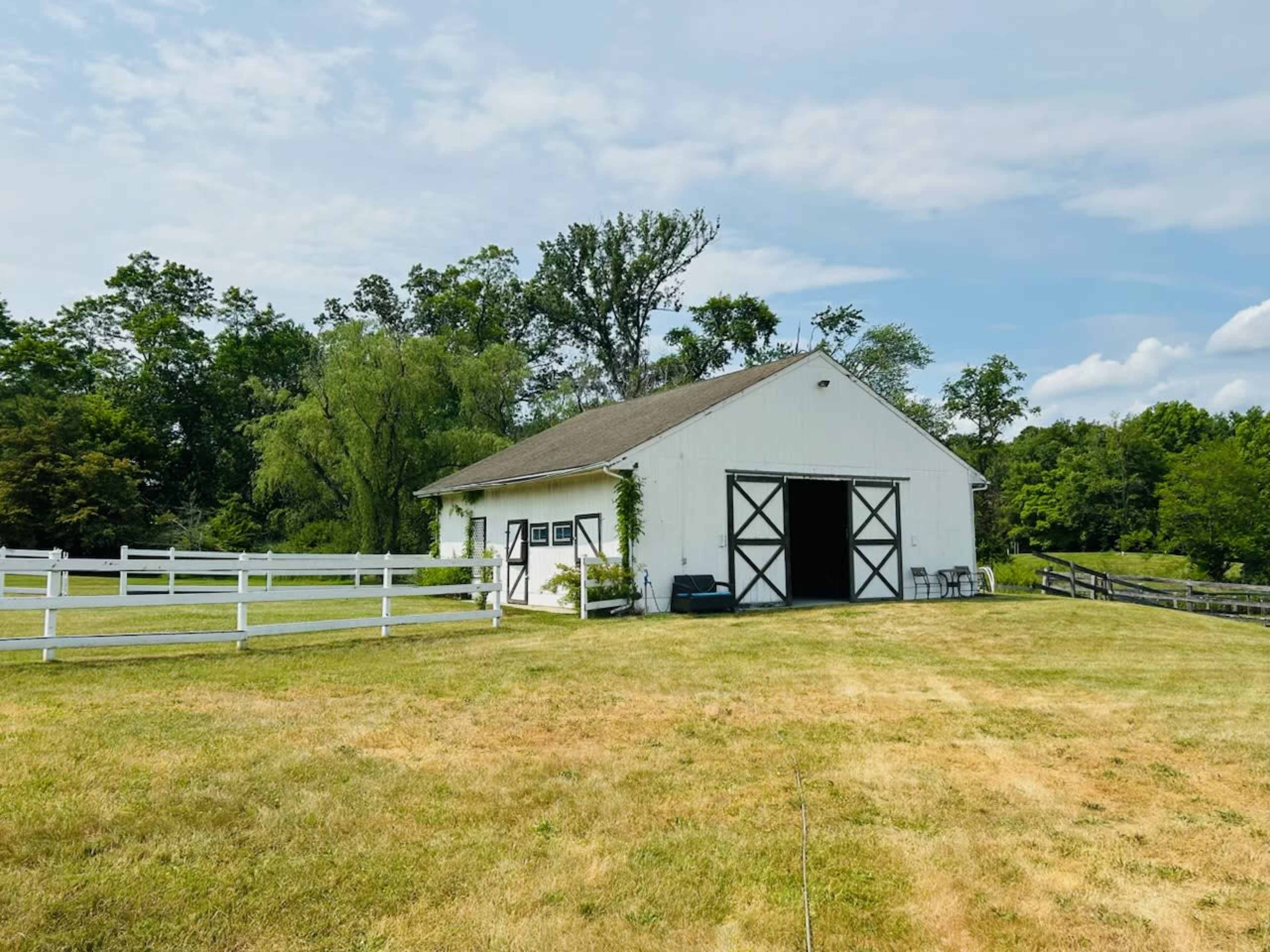 A white barn situated on a grassy field, surrounded by trees and a white fence.