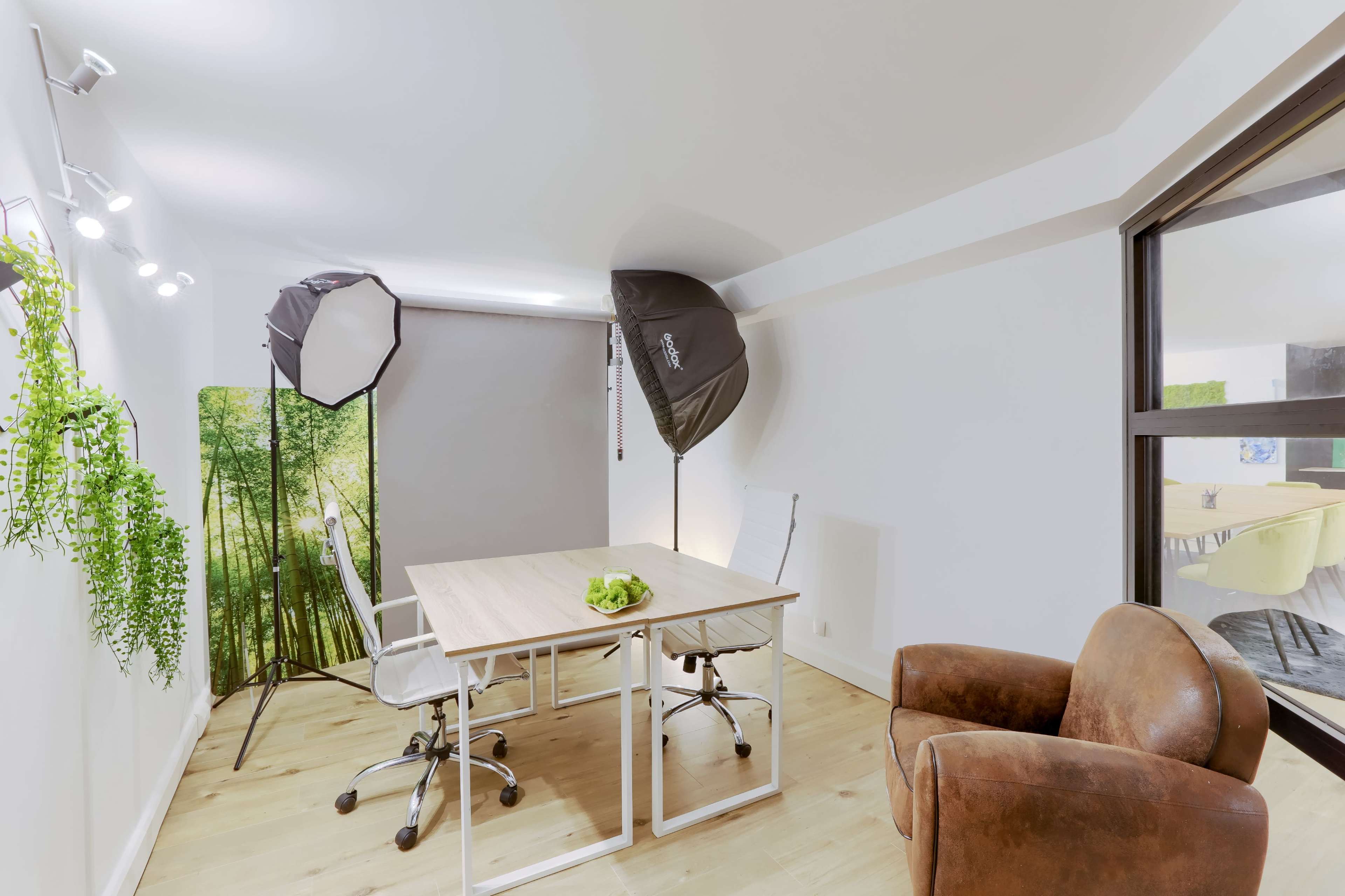 A modern office space features two desks with chairs, a brown armchair, and softbox lights positioned next to a green backdrop.