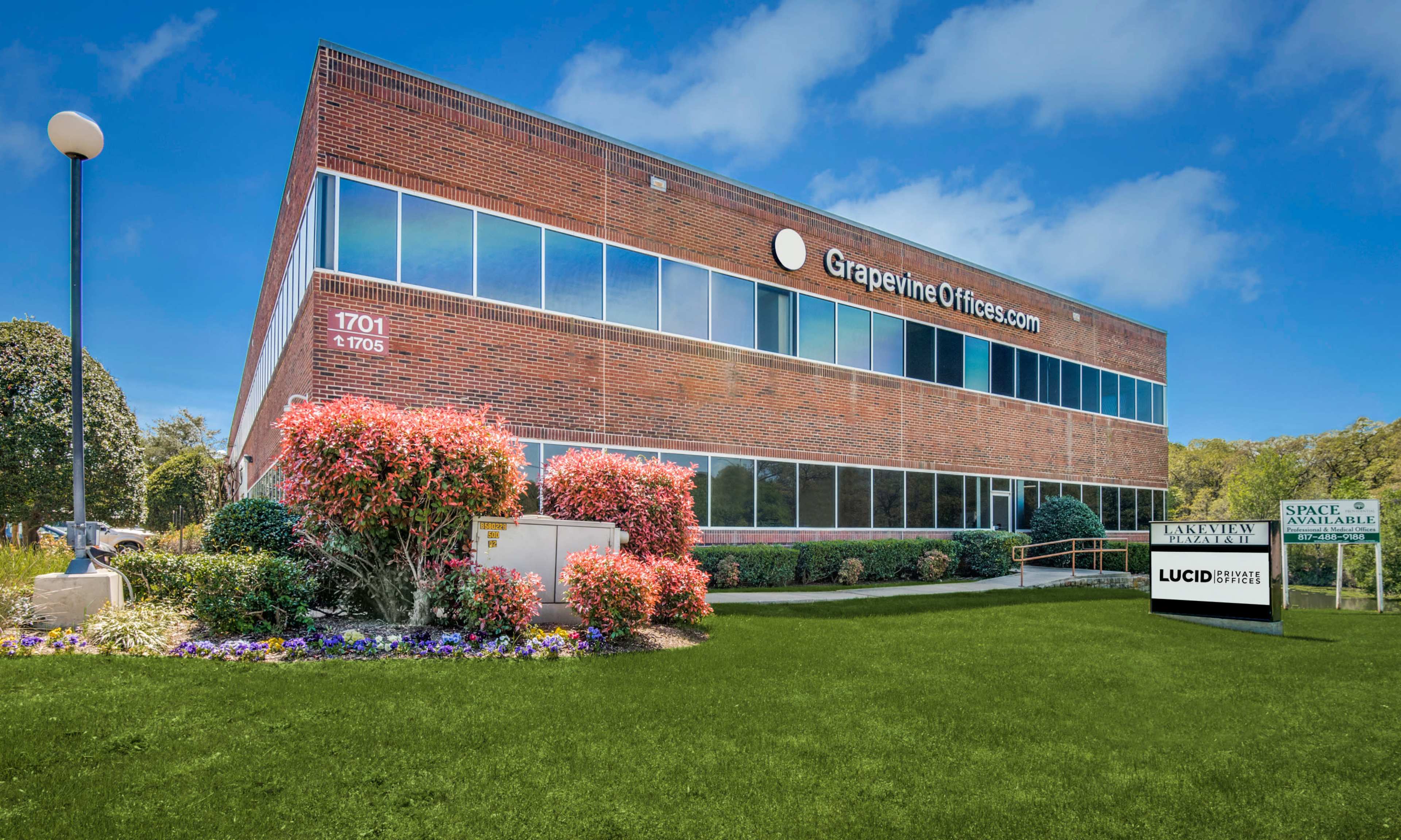 A red-brick office building with large windows and a landscaped front area featuring flowers and a sign displaying available office space.