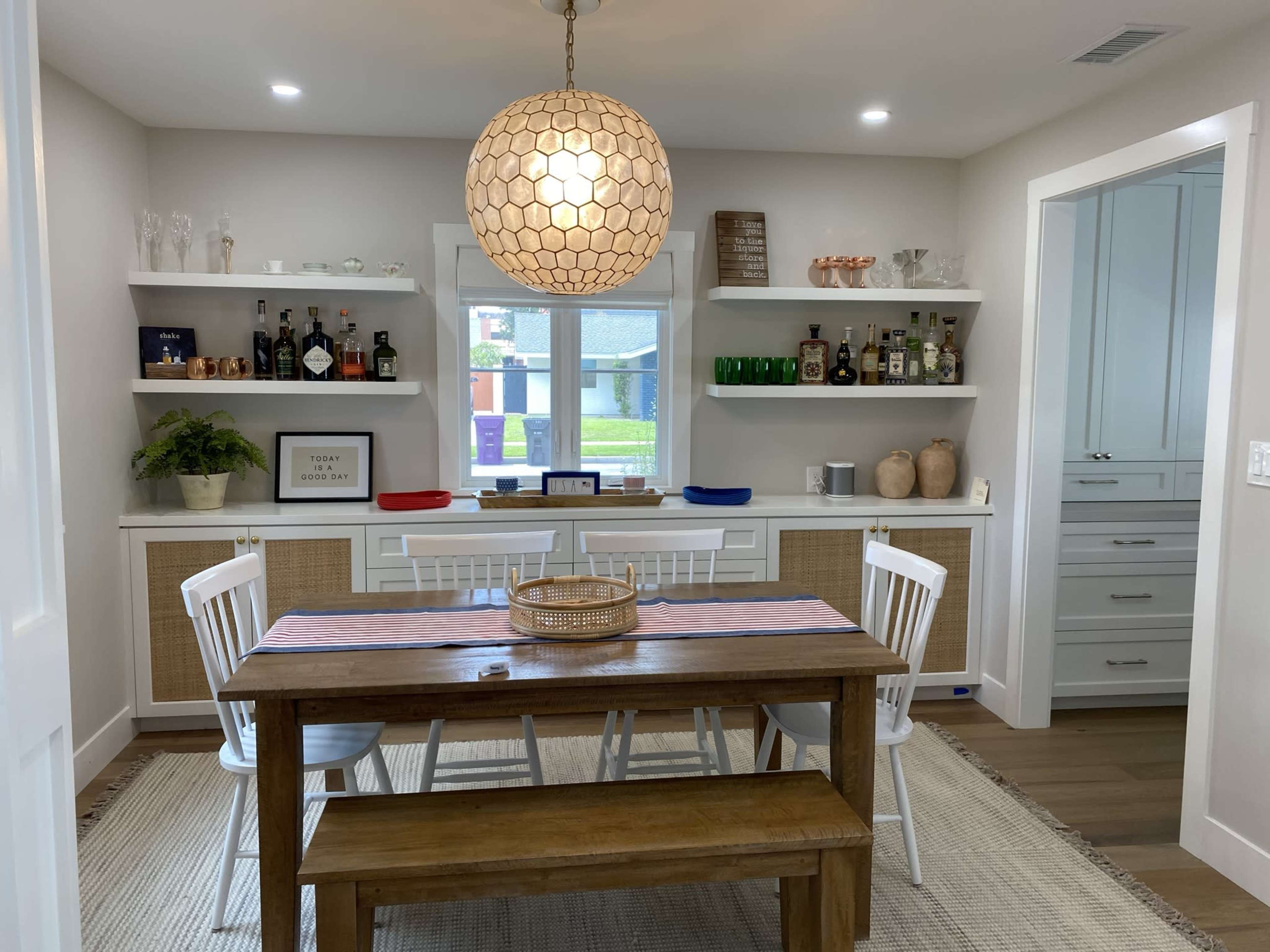 A wooden dining table with white chairs is set under a large pendant light in a room featuring built-in shelves adorned with various bottles and decorative items.