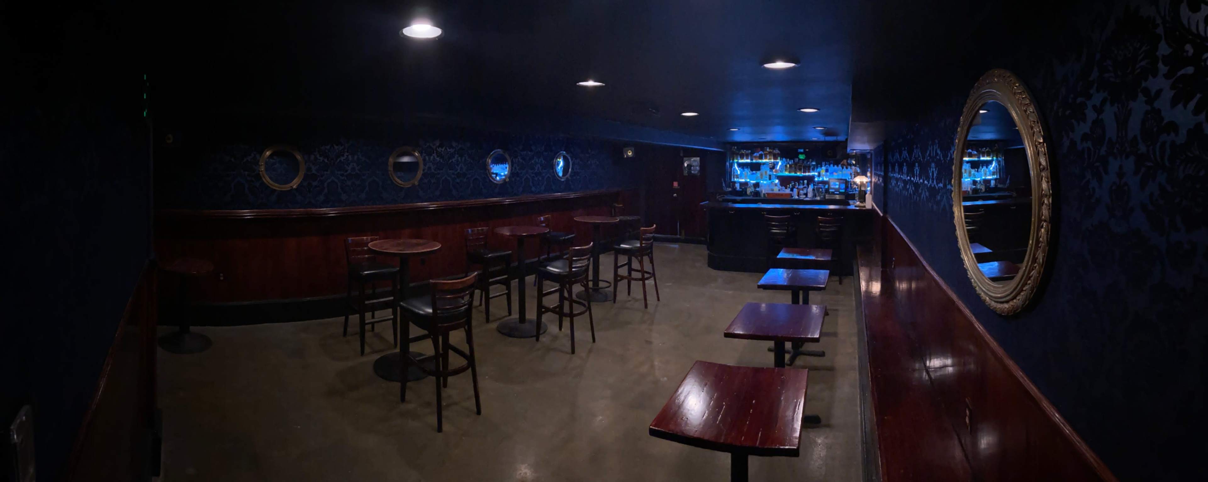 A dimly lit bar interior with wooden tables and chairs arranged around the room, and a bar counter at the far end.