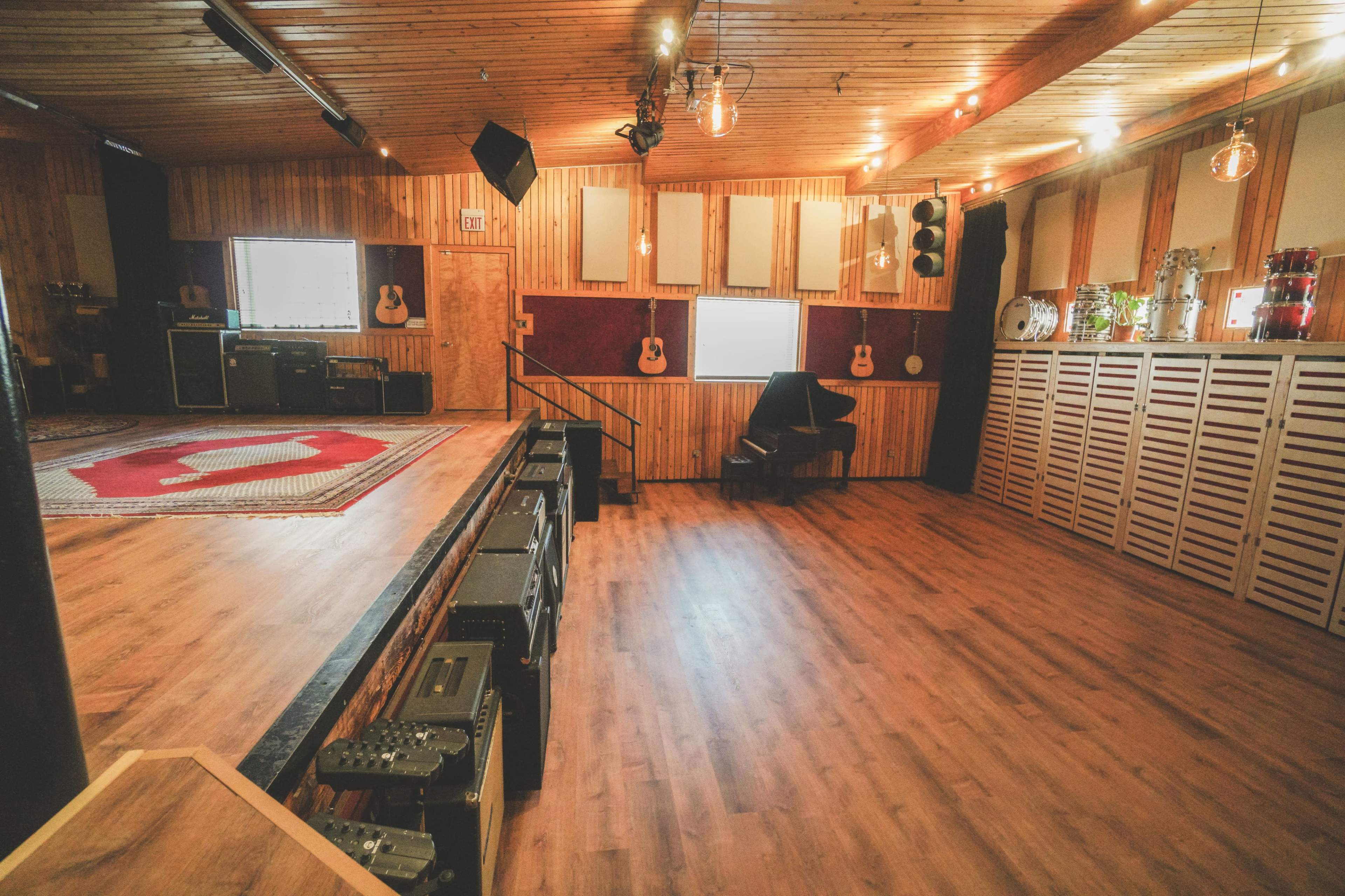 The image shows a music studio with wooden walls, a red rug, amplifiers, and various guitars displayed on shelves.