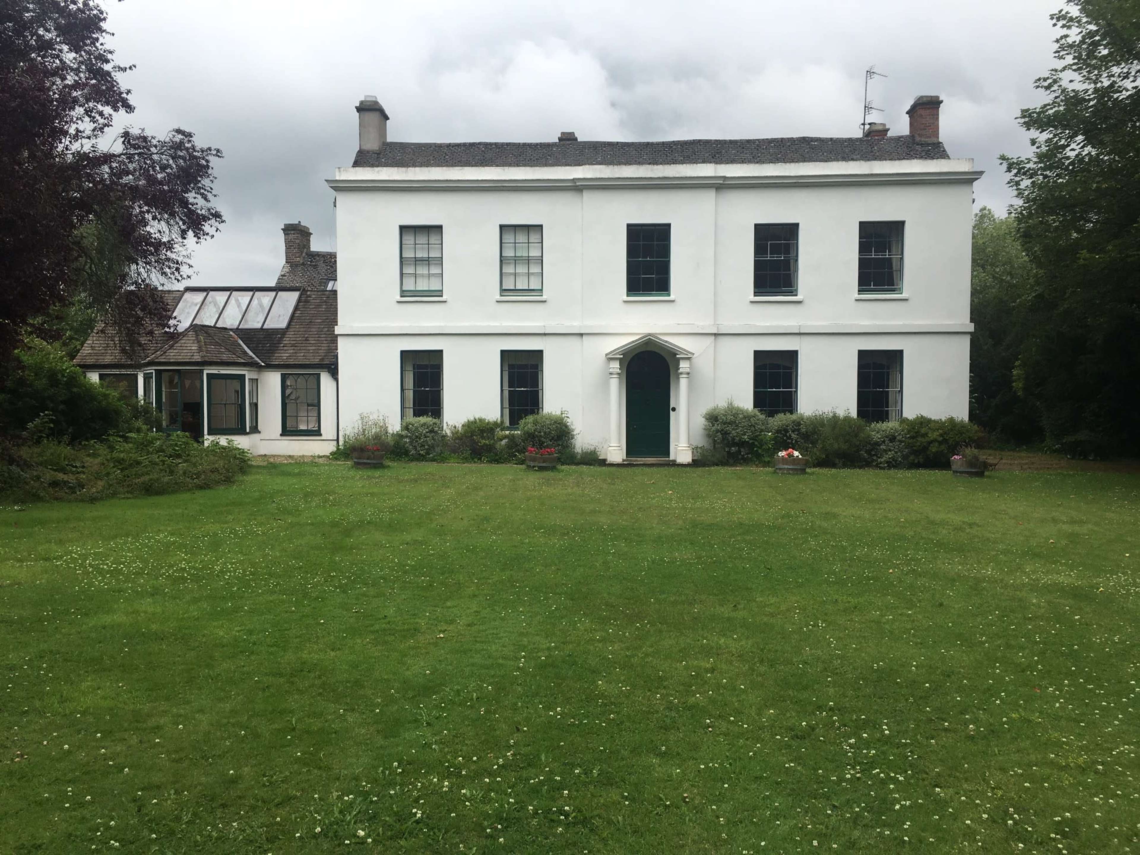 A white two-story house with green windows and a front door is situated on a grassy lawn surrounded by trees and shrubs.