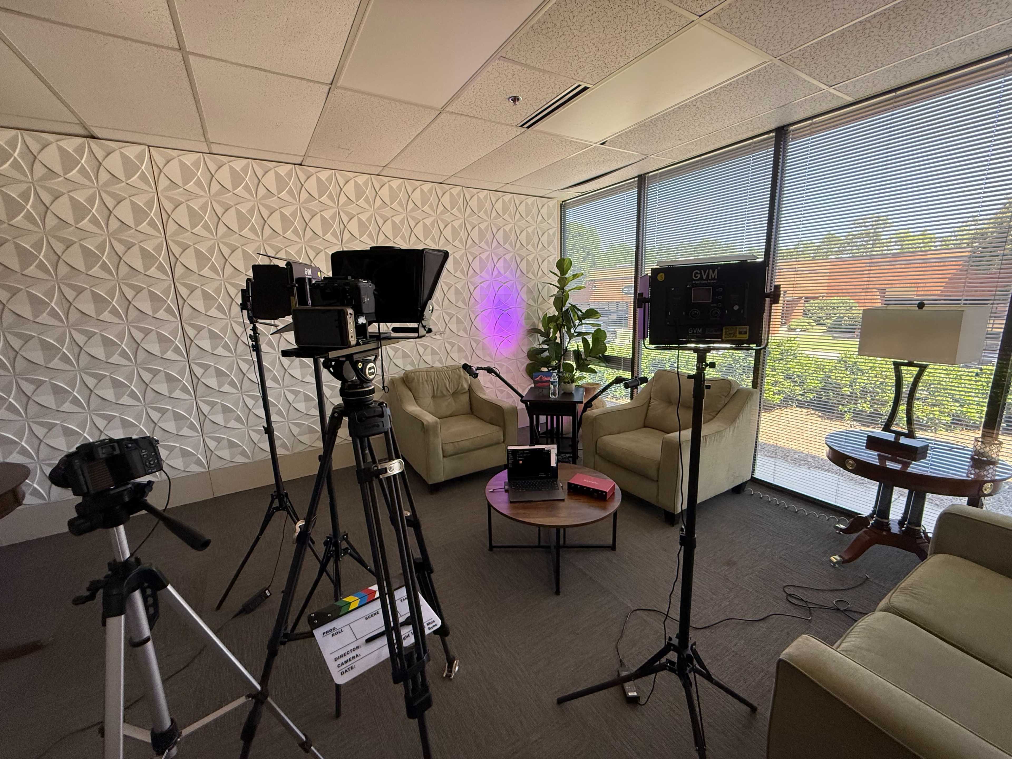 The image shows a well-lit studio setup featuring two beige chairs, a small table, and multiple cameras and lighting equipment arranged in a bright room with large windows.