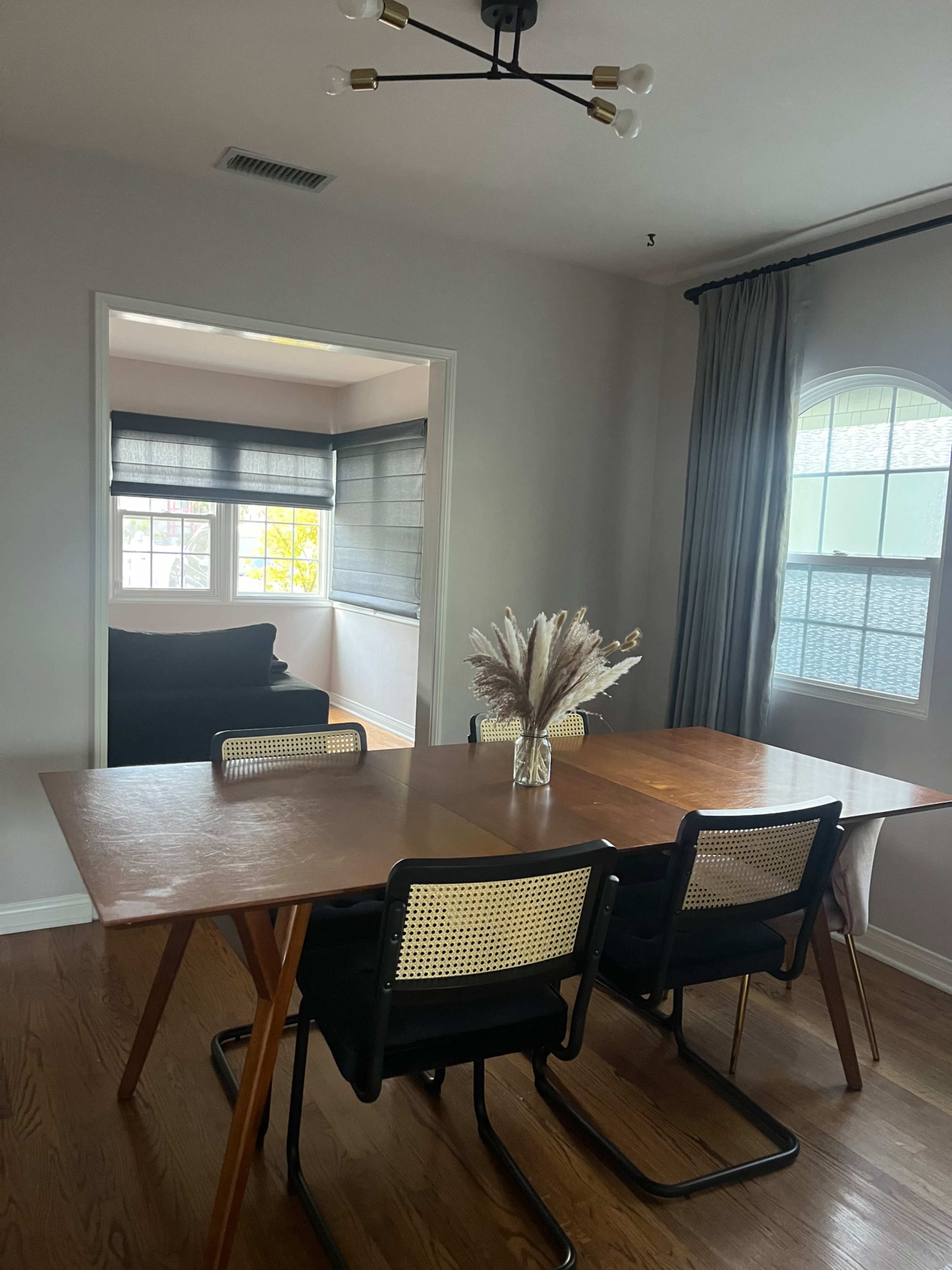 A dining area features a wooden table with black and cane chairs, illuminated by a modern light fixture, adjacent to a room with large windows and a dark sofa.