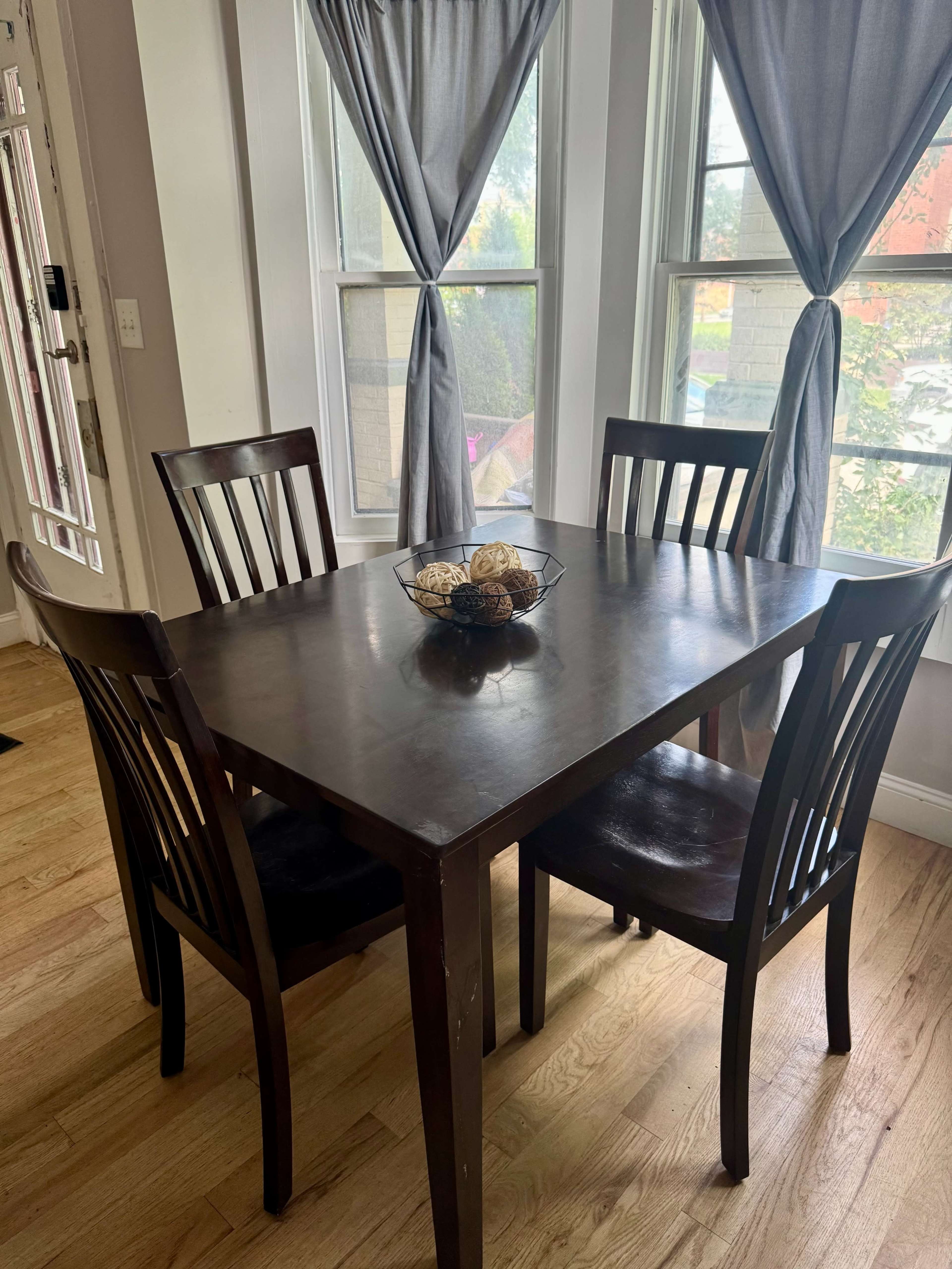 A wooden table with four chairs is set in a brightly lit room with large windows and gray curtains.