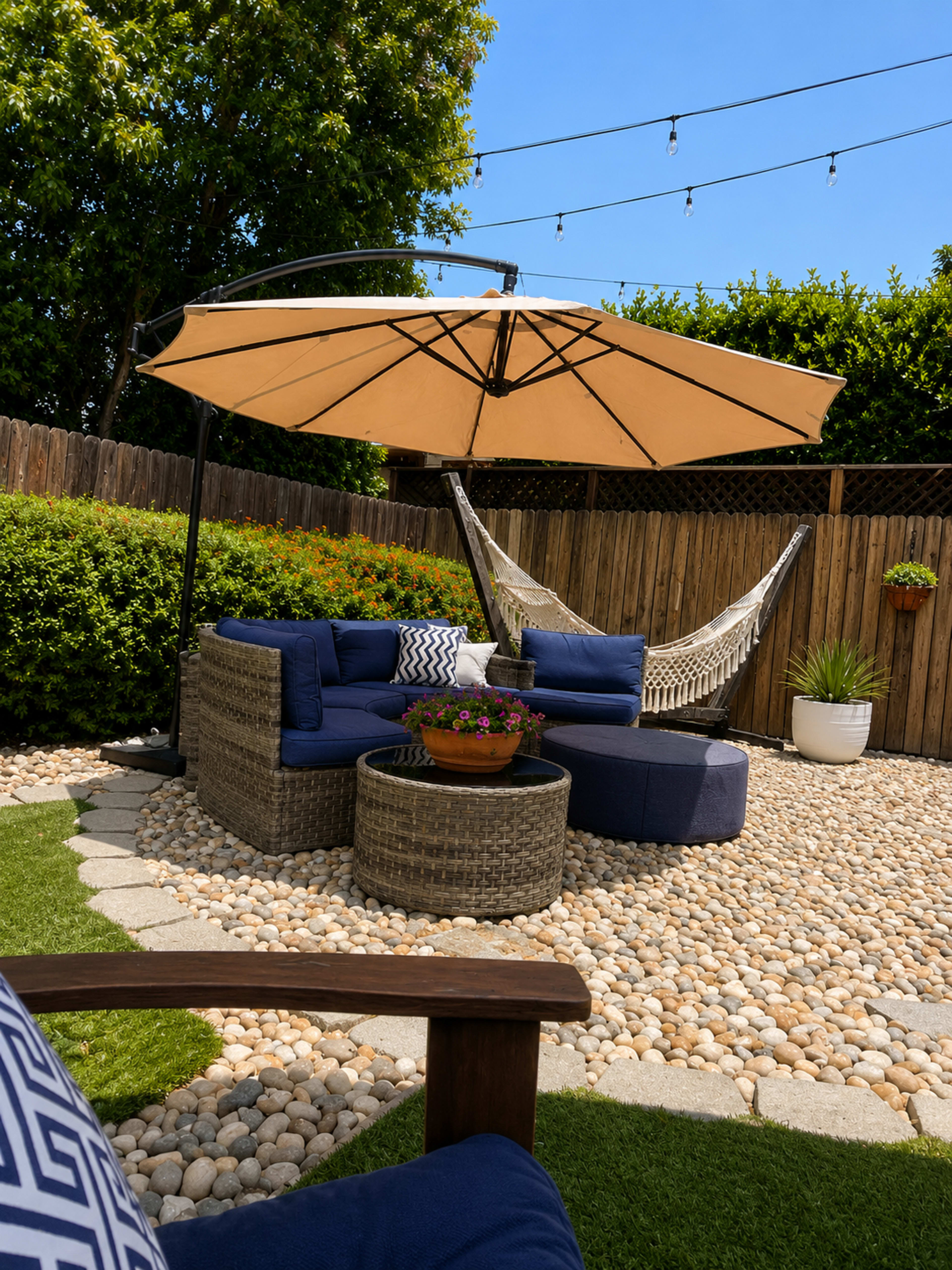The image shows a cozy outdoor seating area with a round table, an umbrella, and a hammock, surrounded by greenery and a stone path.