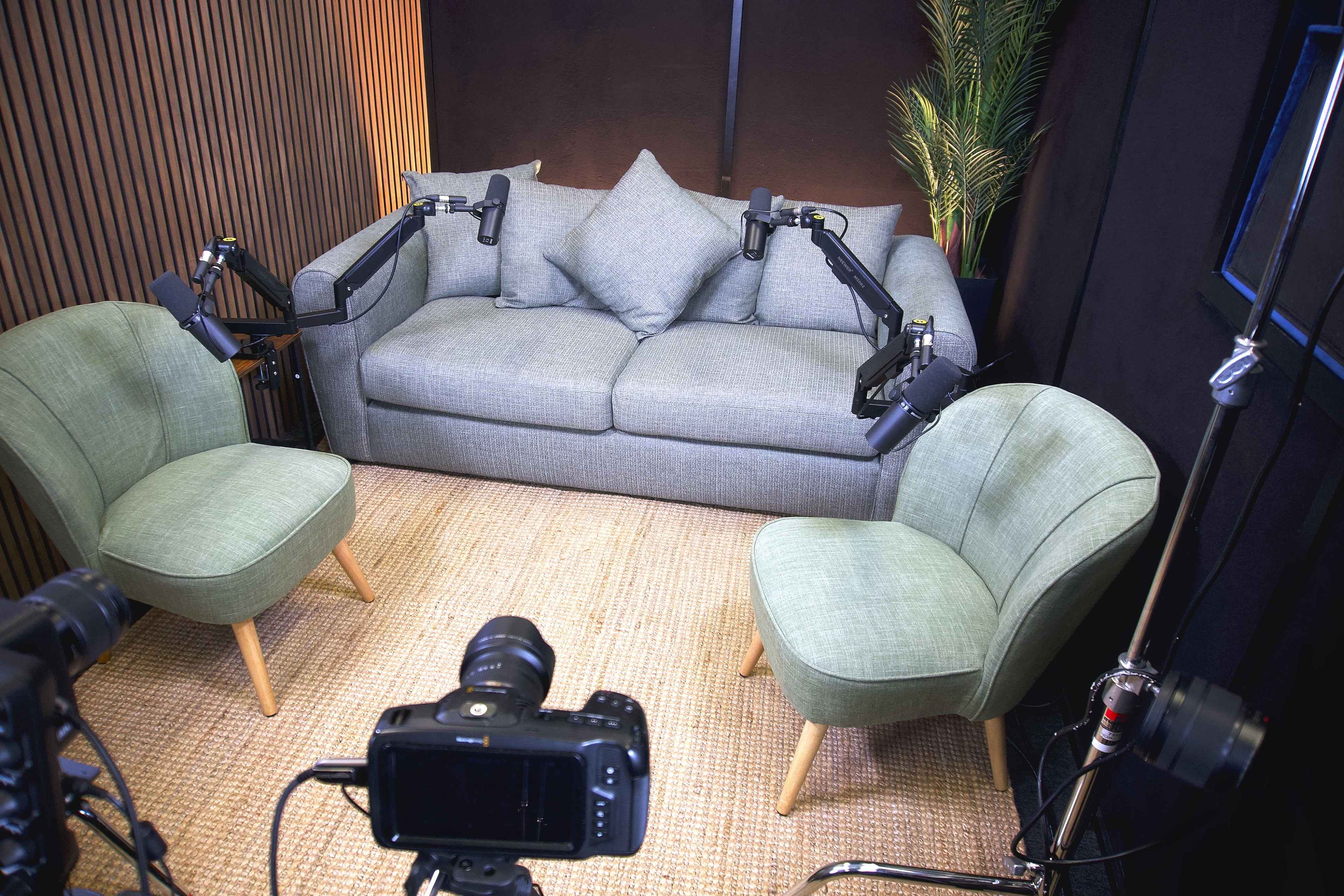 The image shows a small recording studio setup featuring a gray sofa with two green chairs positioned in front, all arranged on a jute rug, with camera equipment aimed at the seating area.