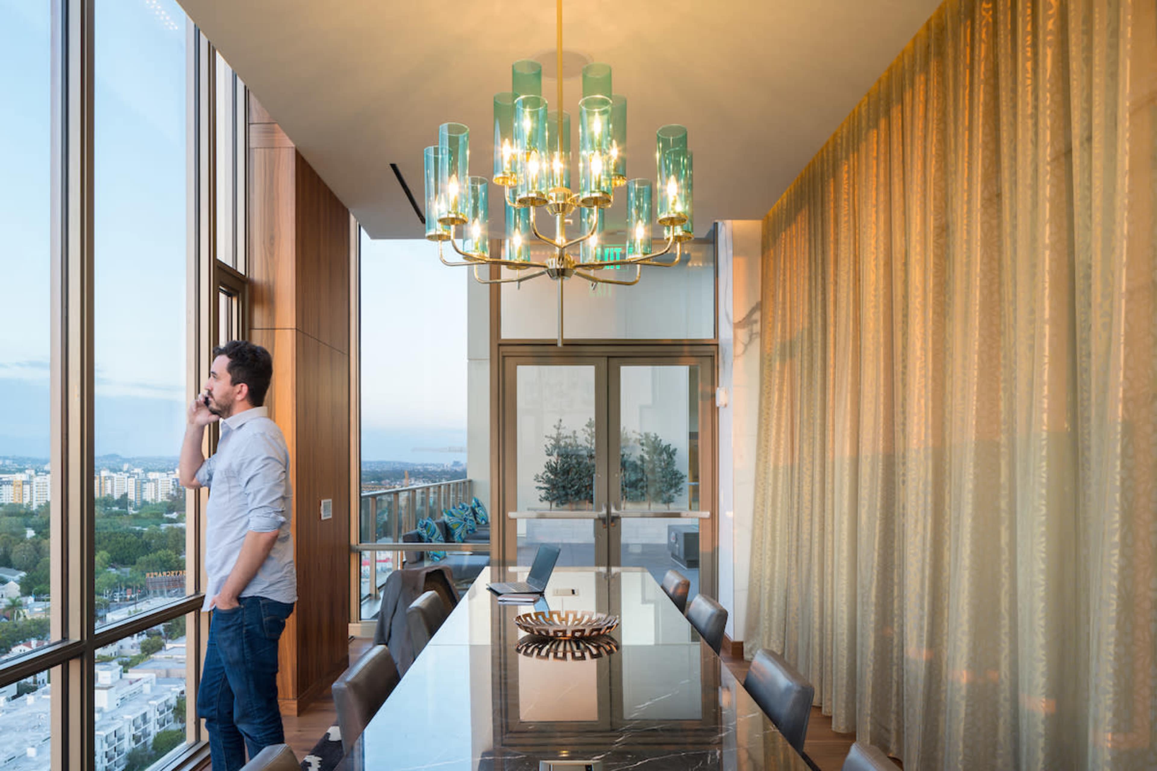 A man is standing by a large window in a modern dining room, holding a phone, with a chandelier and a long table in the foreground.