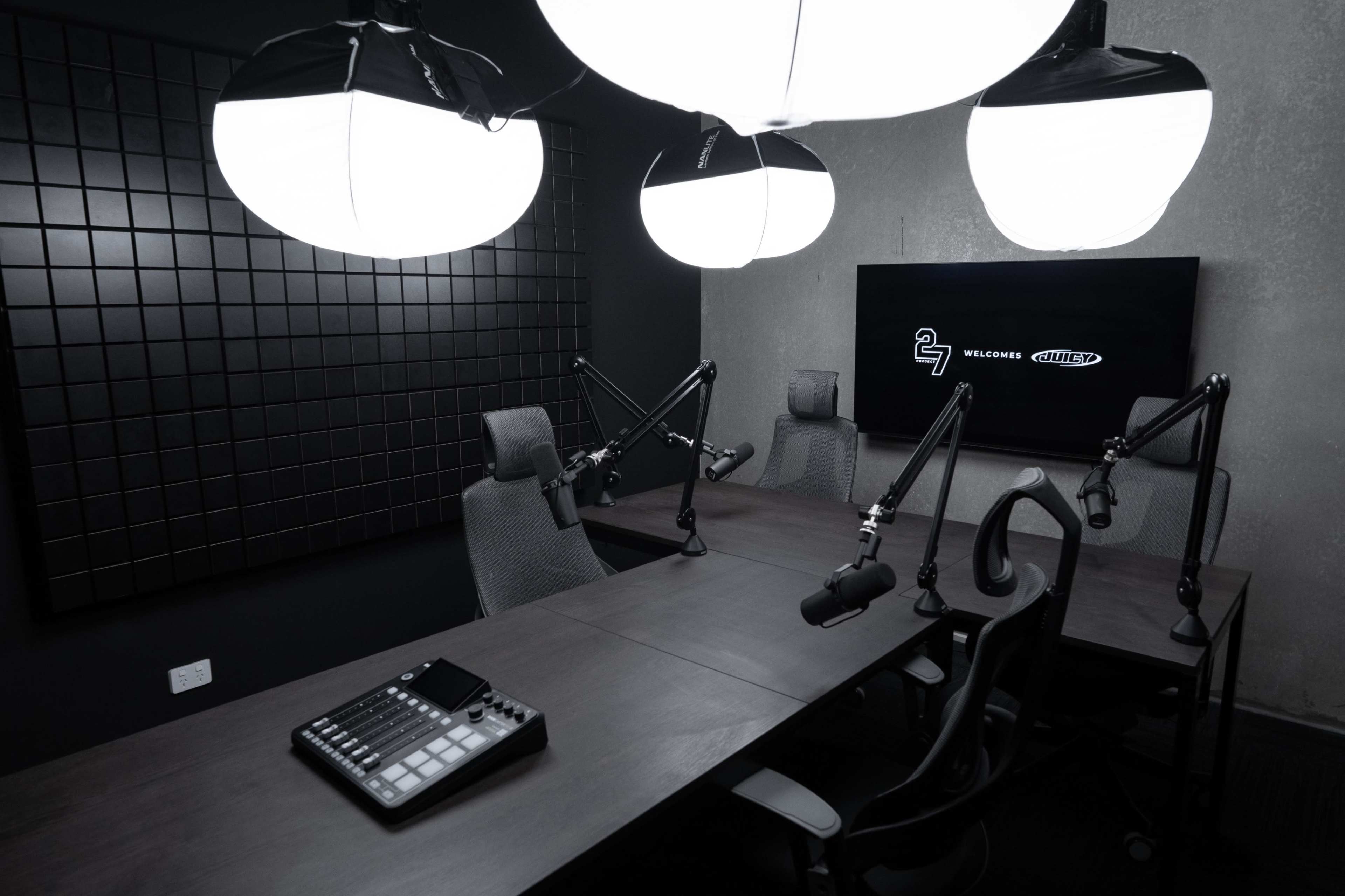 The image shows a modern podcast studio featuring a large wooden table with microphones and an audio mixer, accompanied by three adjustable chairs and dramatic overhead lighting.