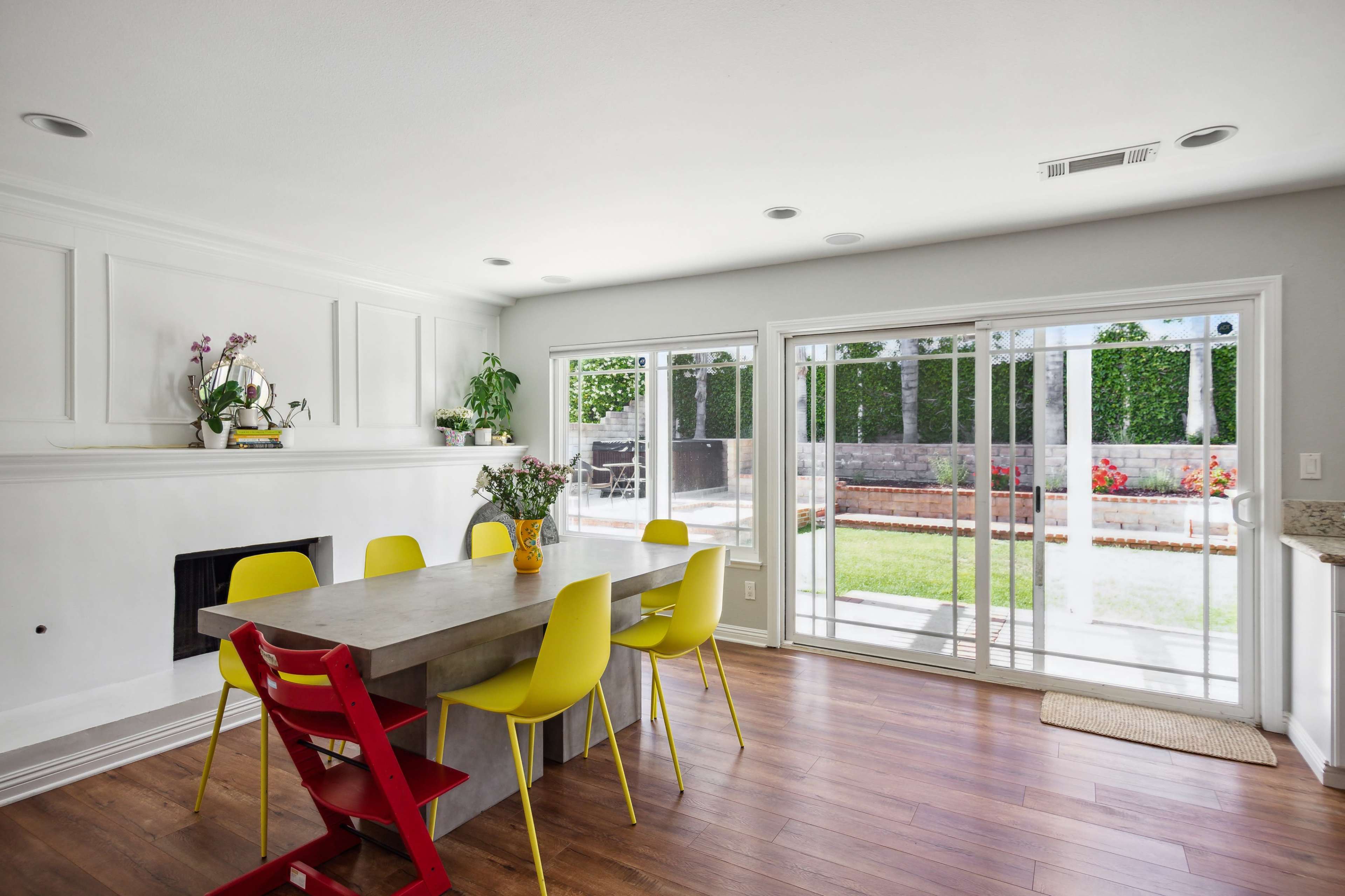 A dining area features a rectangular table surrounded by yellow and red chairs, with a sliding glass door leading to a backyard.