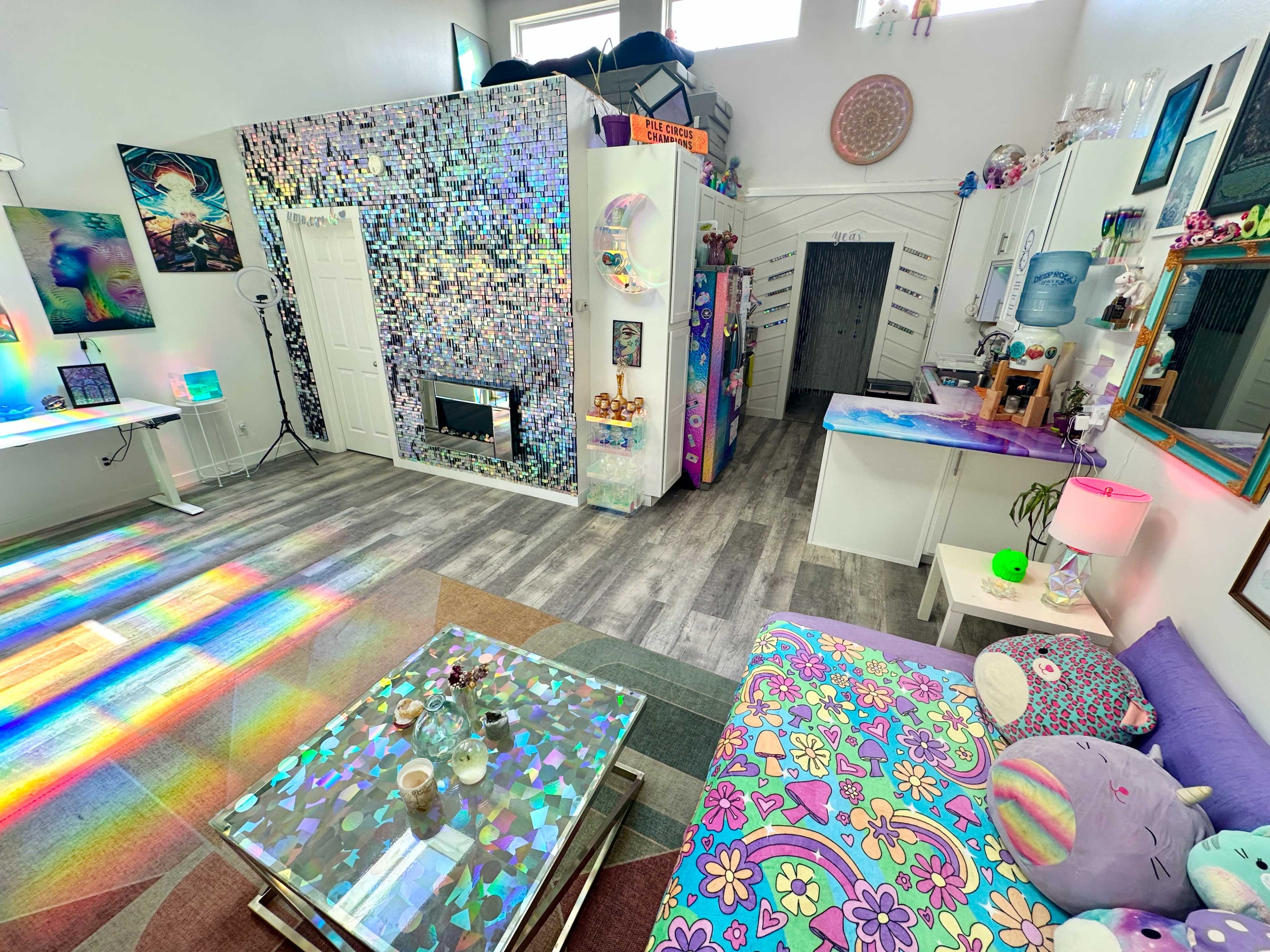 The image shows a brightly lit, colorful living space featuring a sofa with vibrant patterns, a glass coffee table, and a wall adorned with reflective tiles.