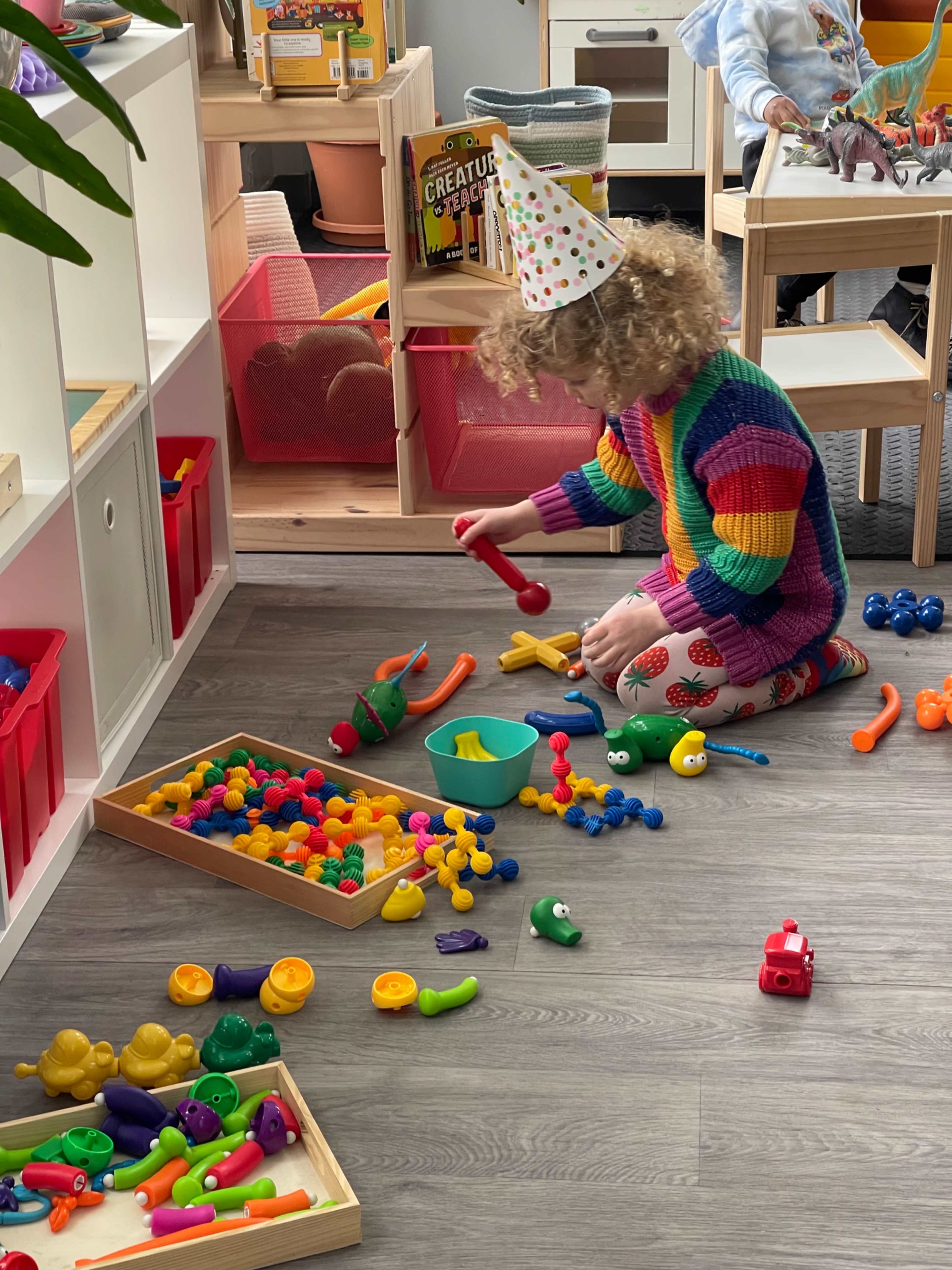 A child wearing a party hat plays with colorful toys on the floor of a playroom.