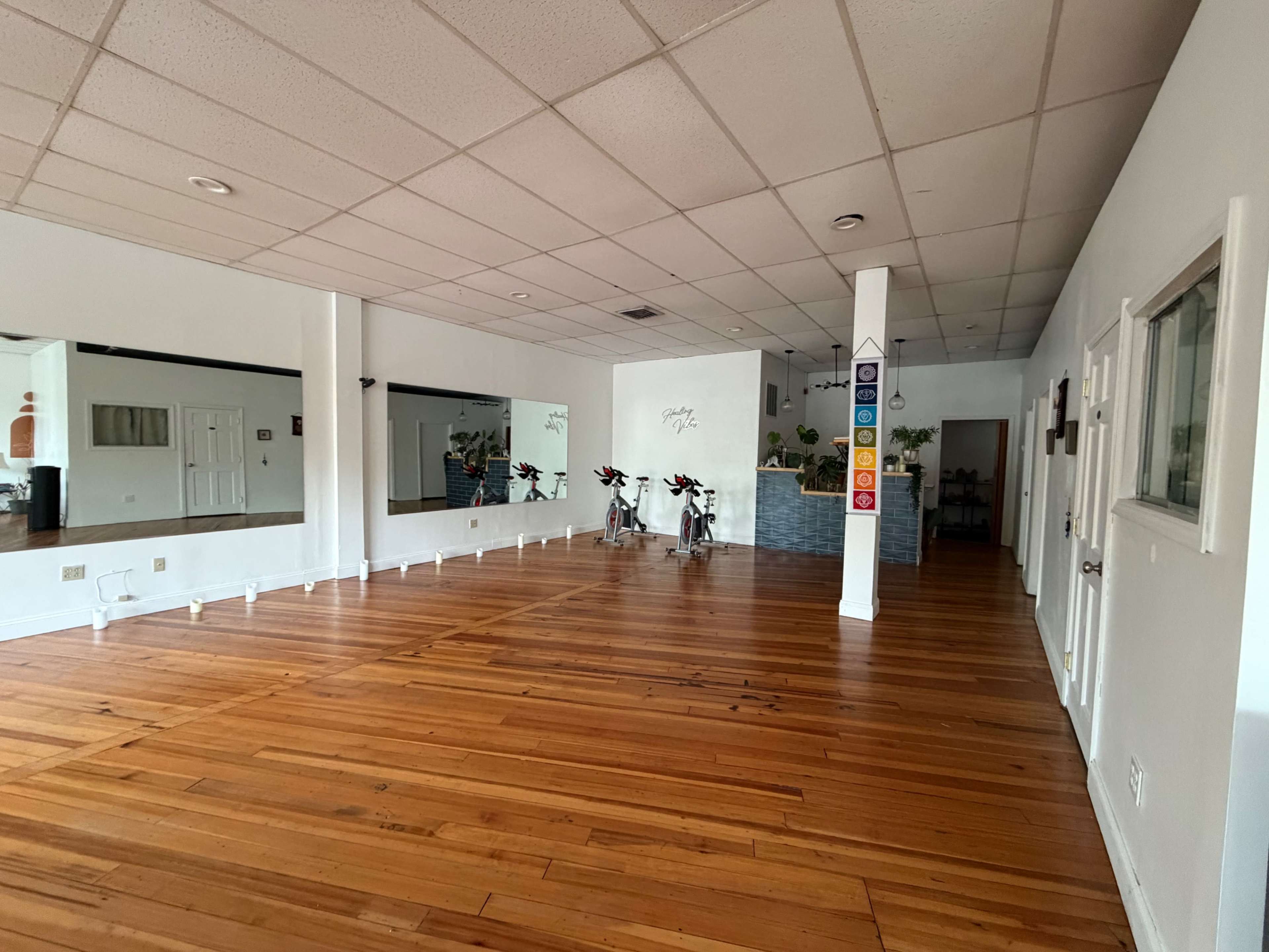 The image shows an empty fitness studio with wooden flooring, large mirrors on one wall, and exercise bikes positioned near a partition.