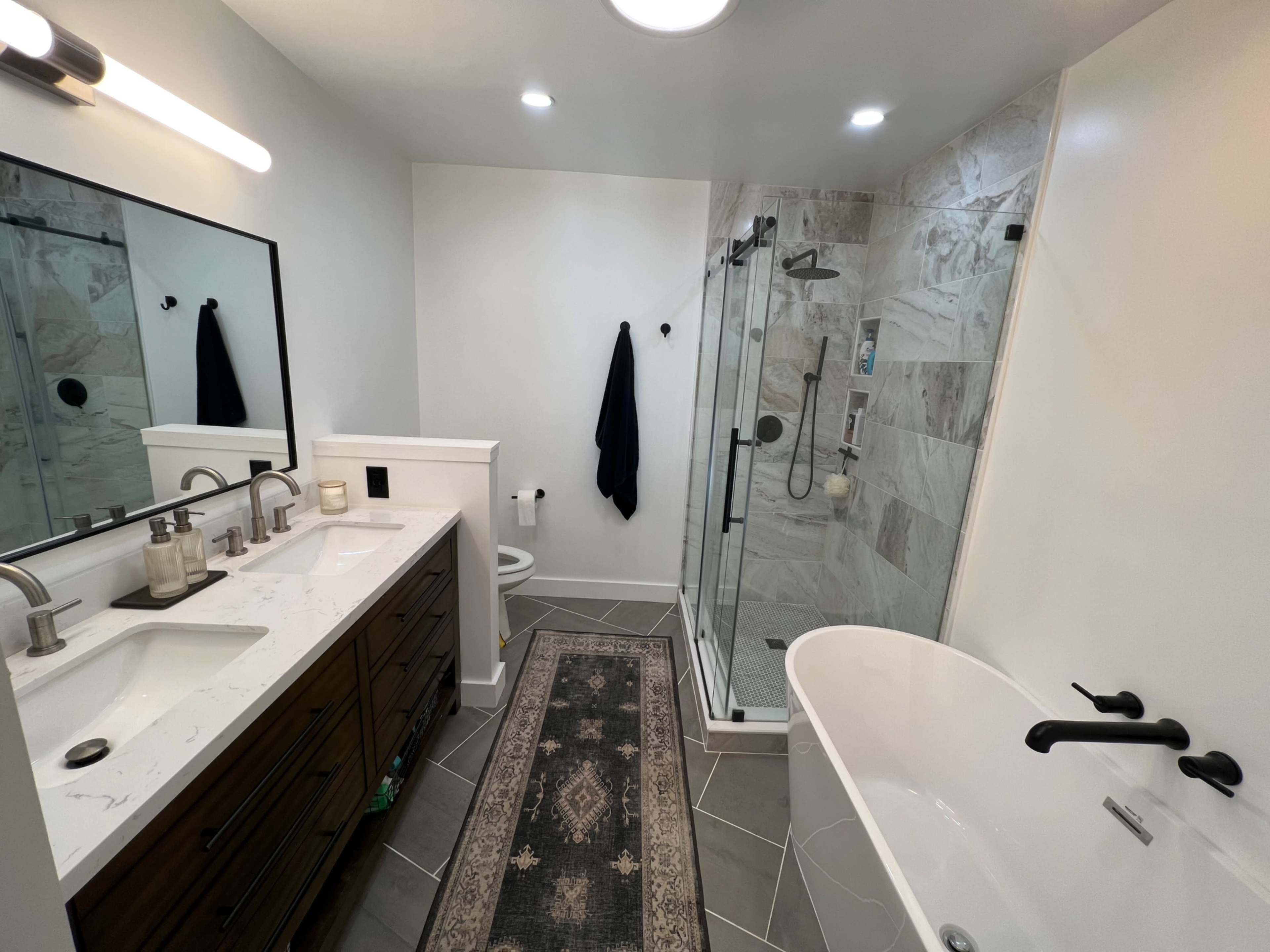 The image shows a modern bathroom featuring a double sink vanity, a walk-in shower, a freestanding bathtub, and a decorative rug on the tiled floor.