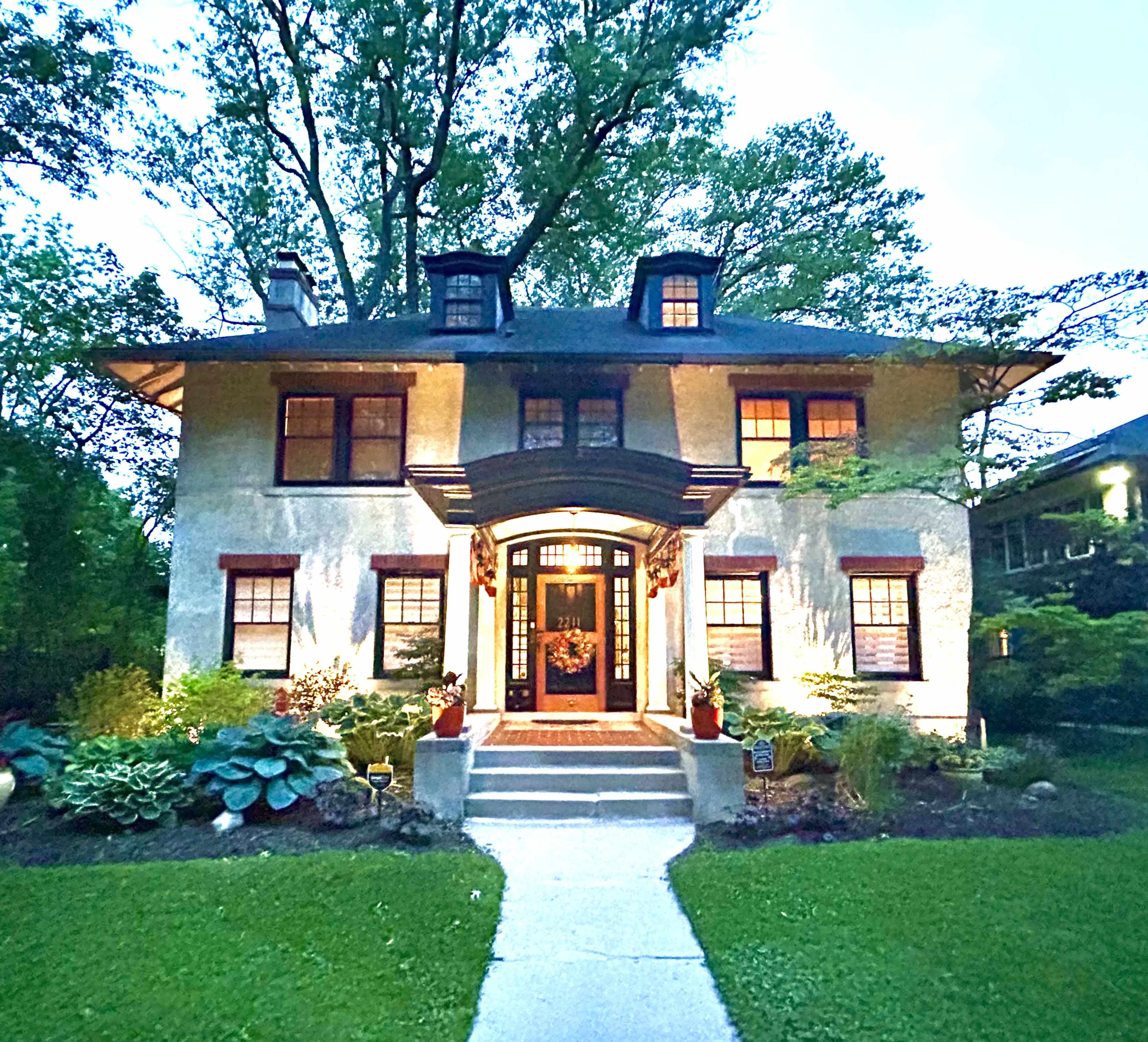 A well-lit, two-story house features a central entrance with a decorative overhang, surrounded by landscaped greenery.