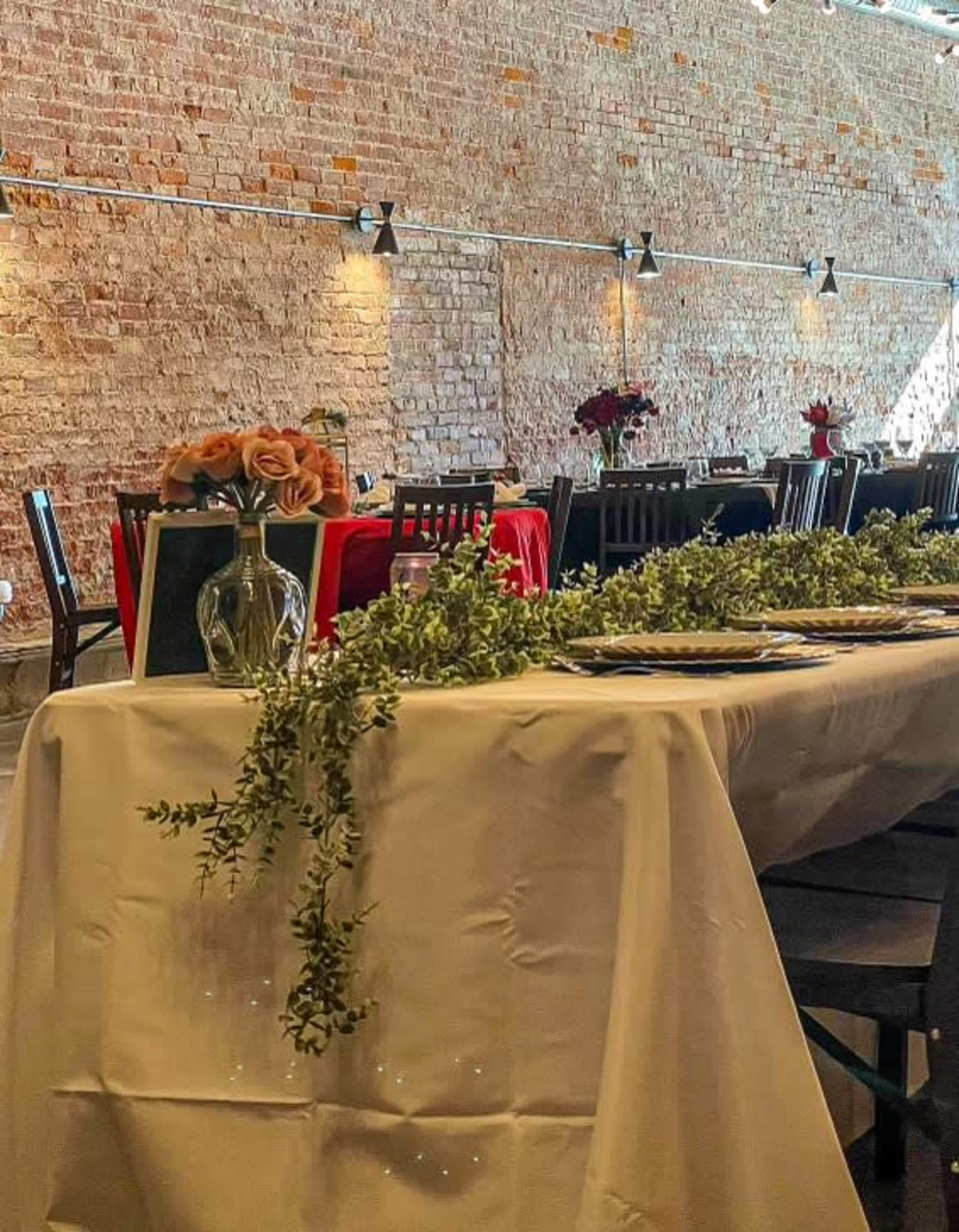 A long dining table is set with floral arrangements and greenery, against a backdrop of a brick wall and additional tables in the distance.