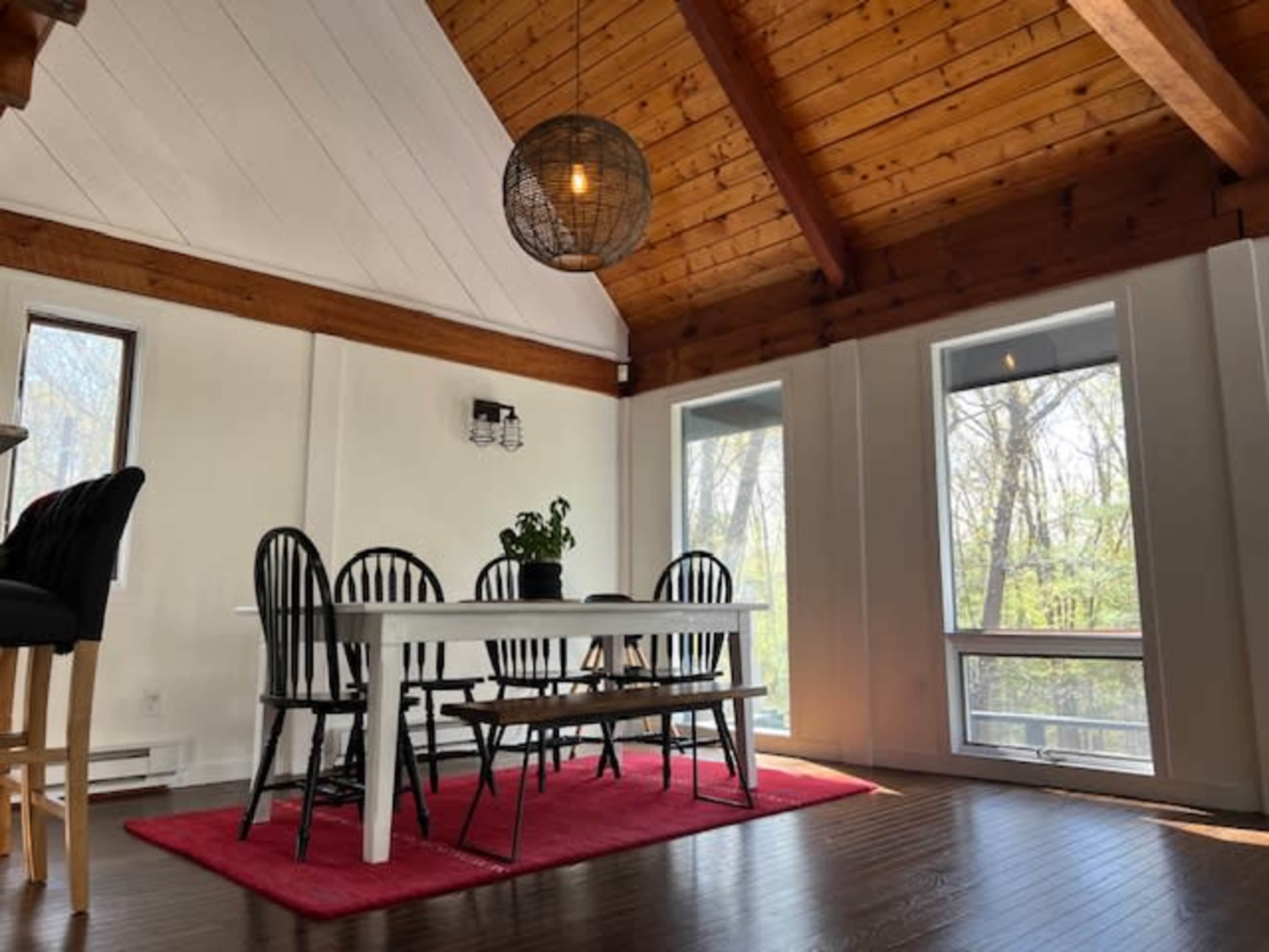 A dining area features a white table surrounded by black chairs, with large windows allowing natural light to enter, and a red rug on the wooden floor.