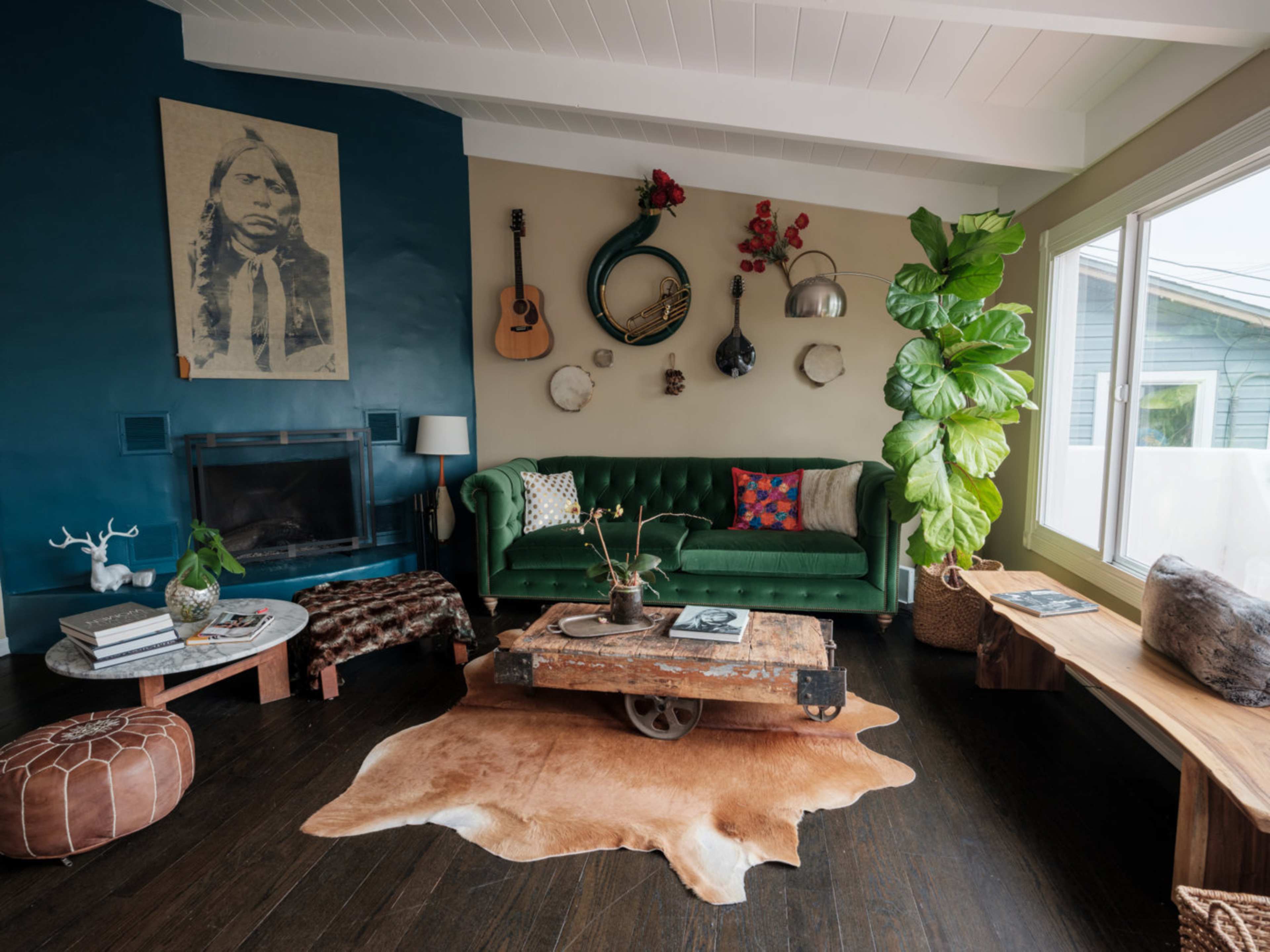 The room features a green sofa, a wooden coffee table on wheels, various musical instruments hanging on the wall, and large plants near a window.