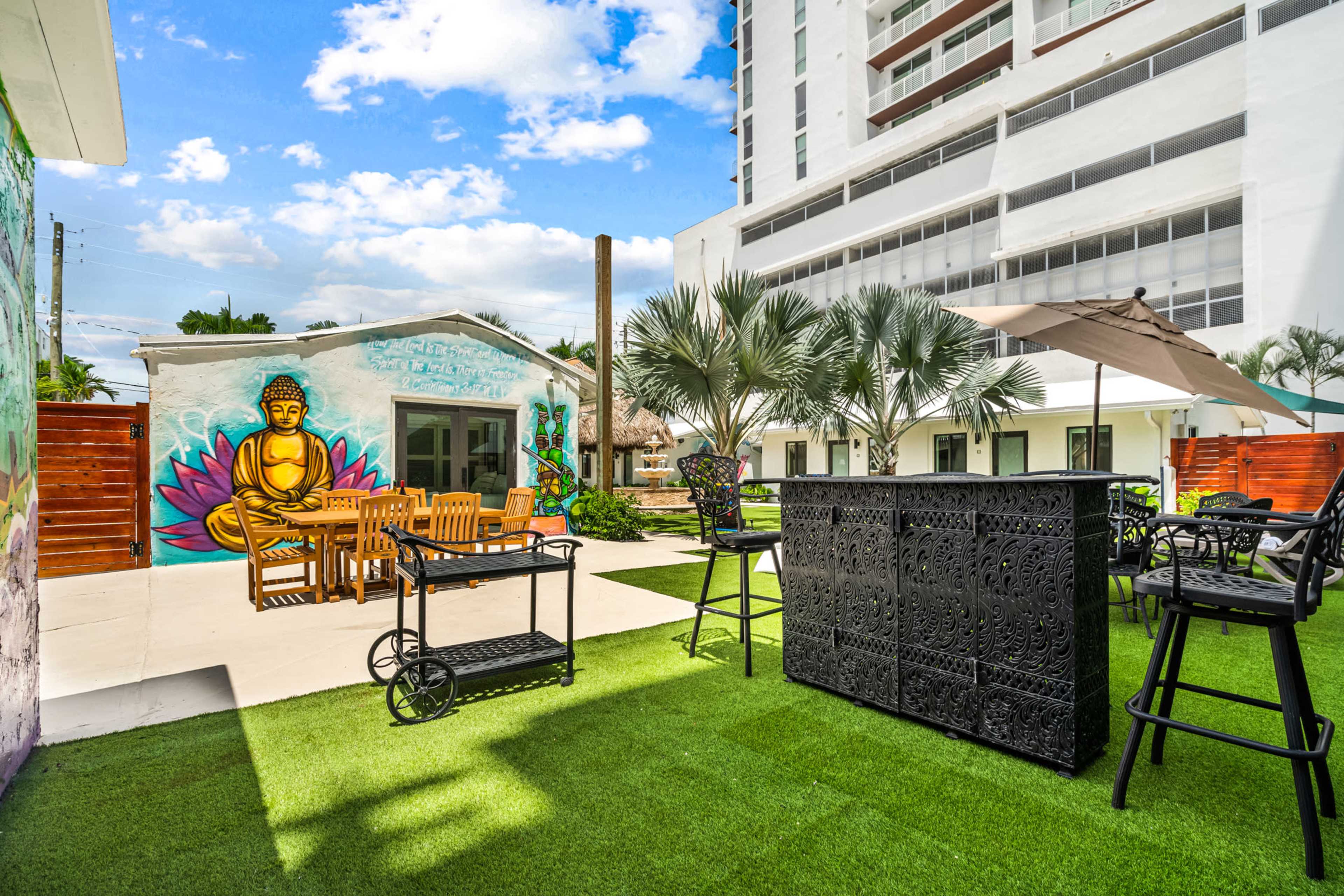 The image shows an outdoor area with seating, a bar, and a mural of a Buddha beside a building, surrounded by palm trees and grass.