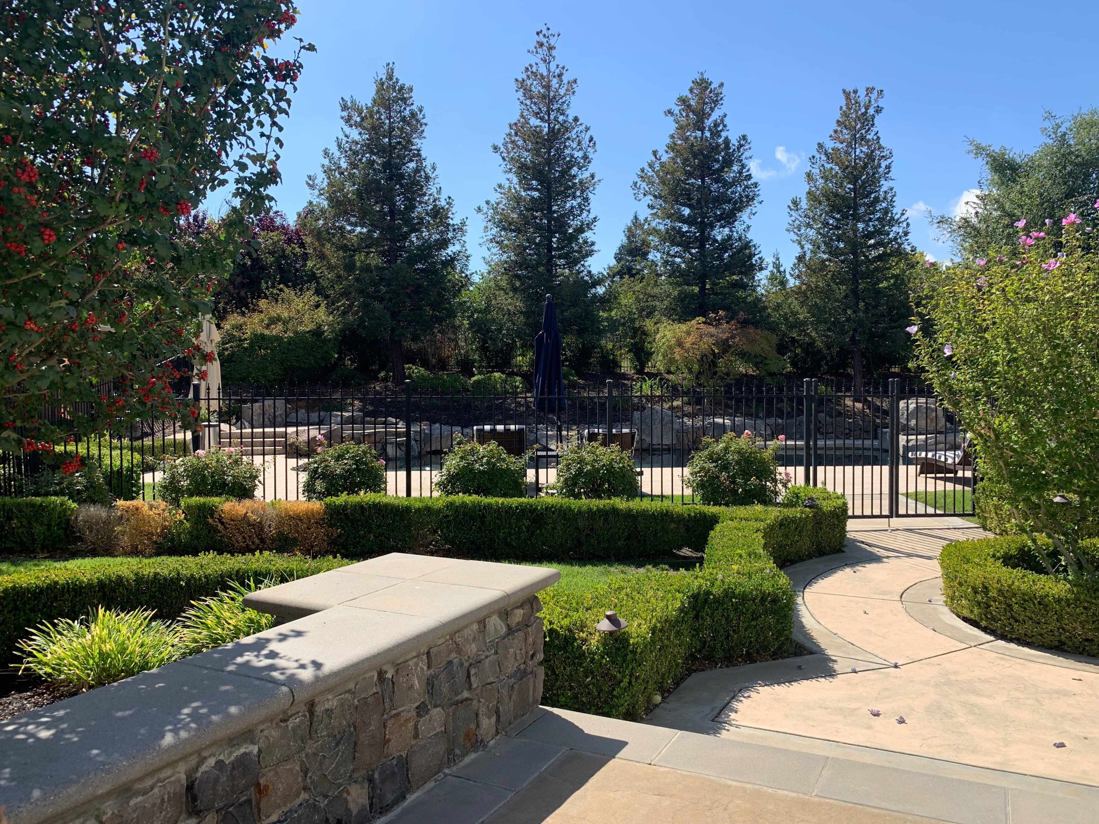 A landscaped garden with neatly trimmed hedges, stone pathways, and a fenced pool area surrounded by trees and flowering plants.