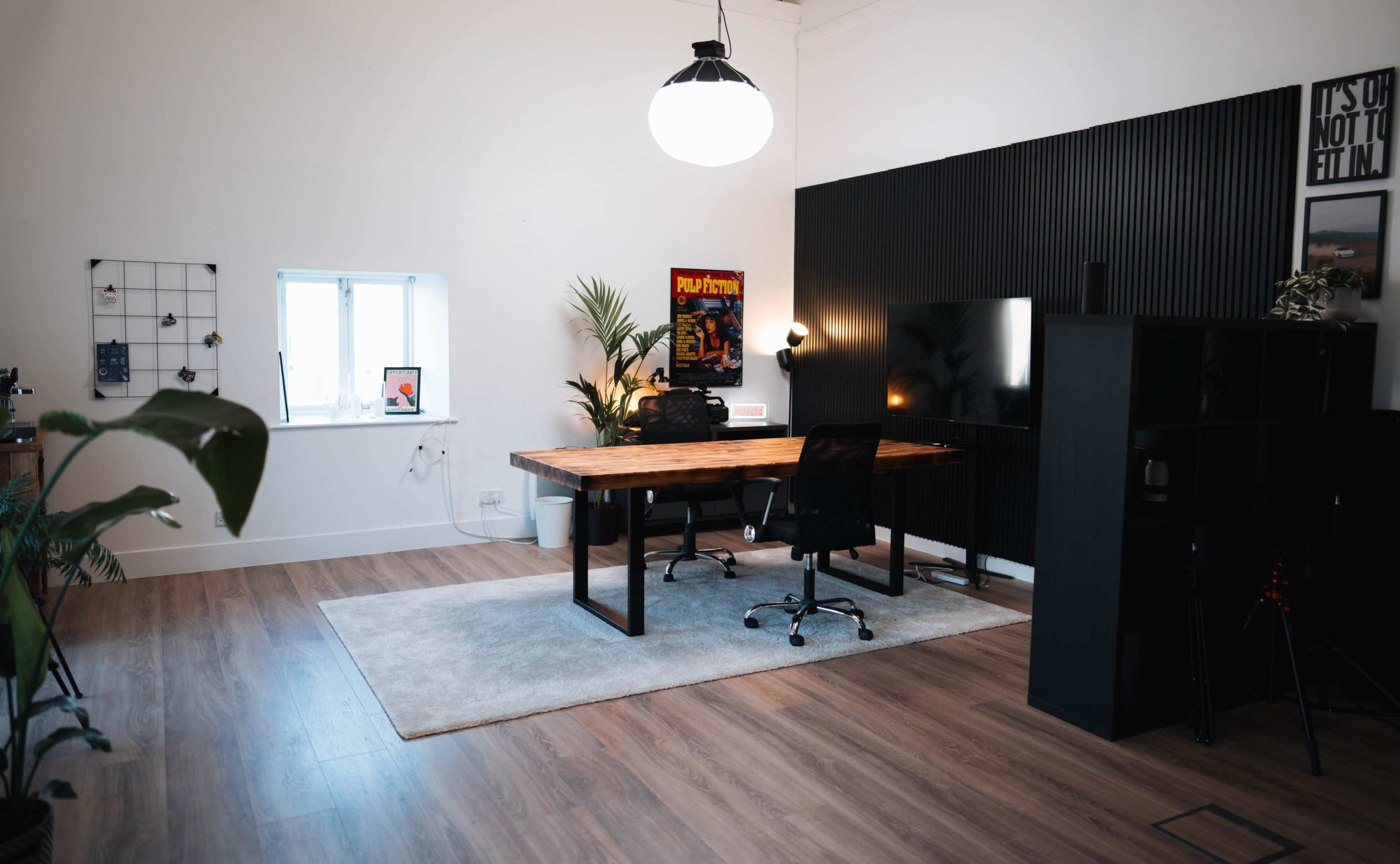 The image shows a modern office space with a large wooden desk, a black chair, a wall-mounted TV, and a plant in the corner.