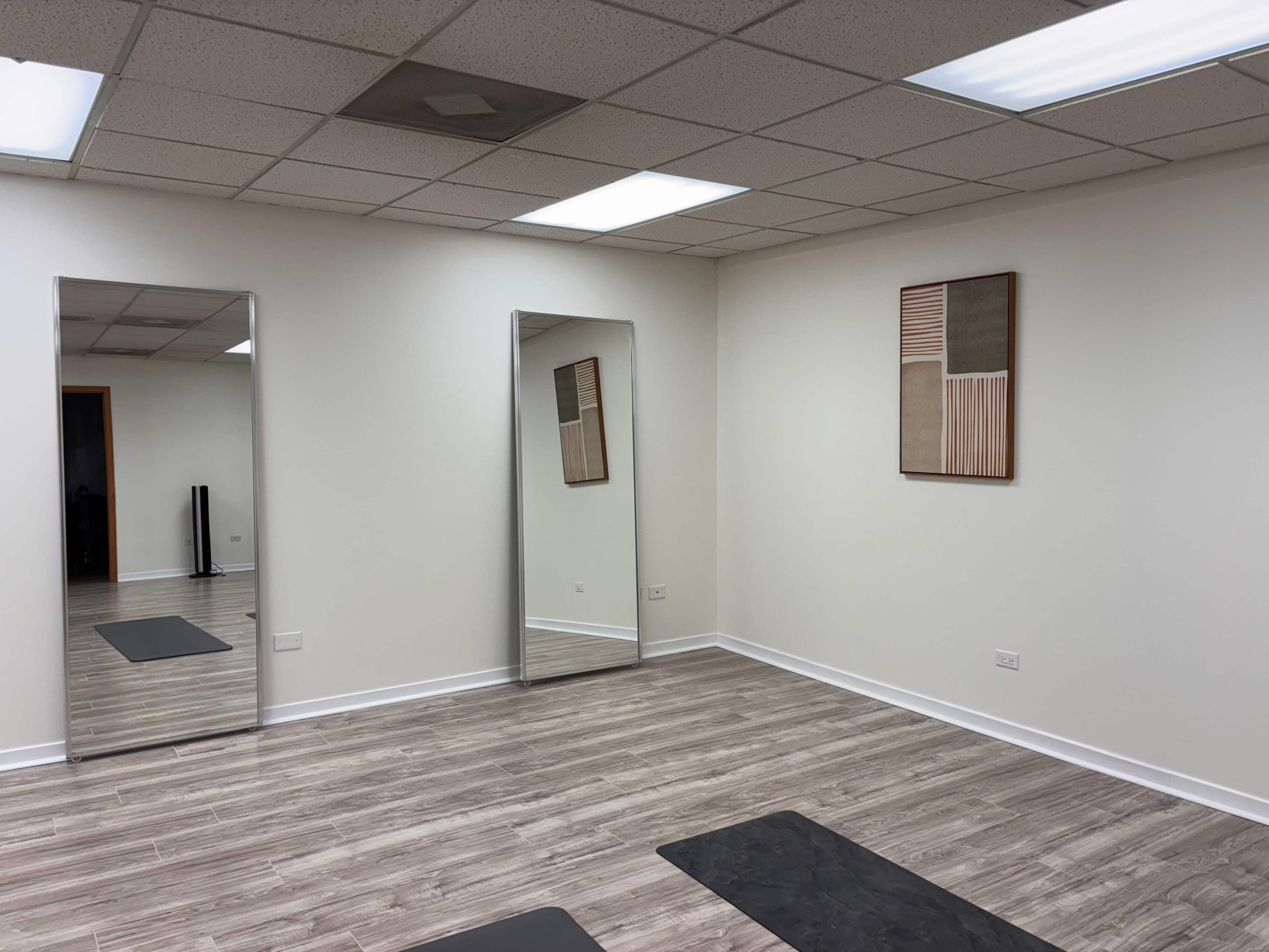 The image shows an empty room with three mirrors, wooden flooring, and a simple wall decoration.