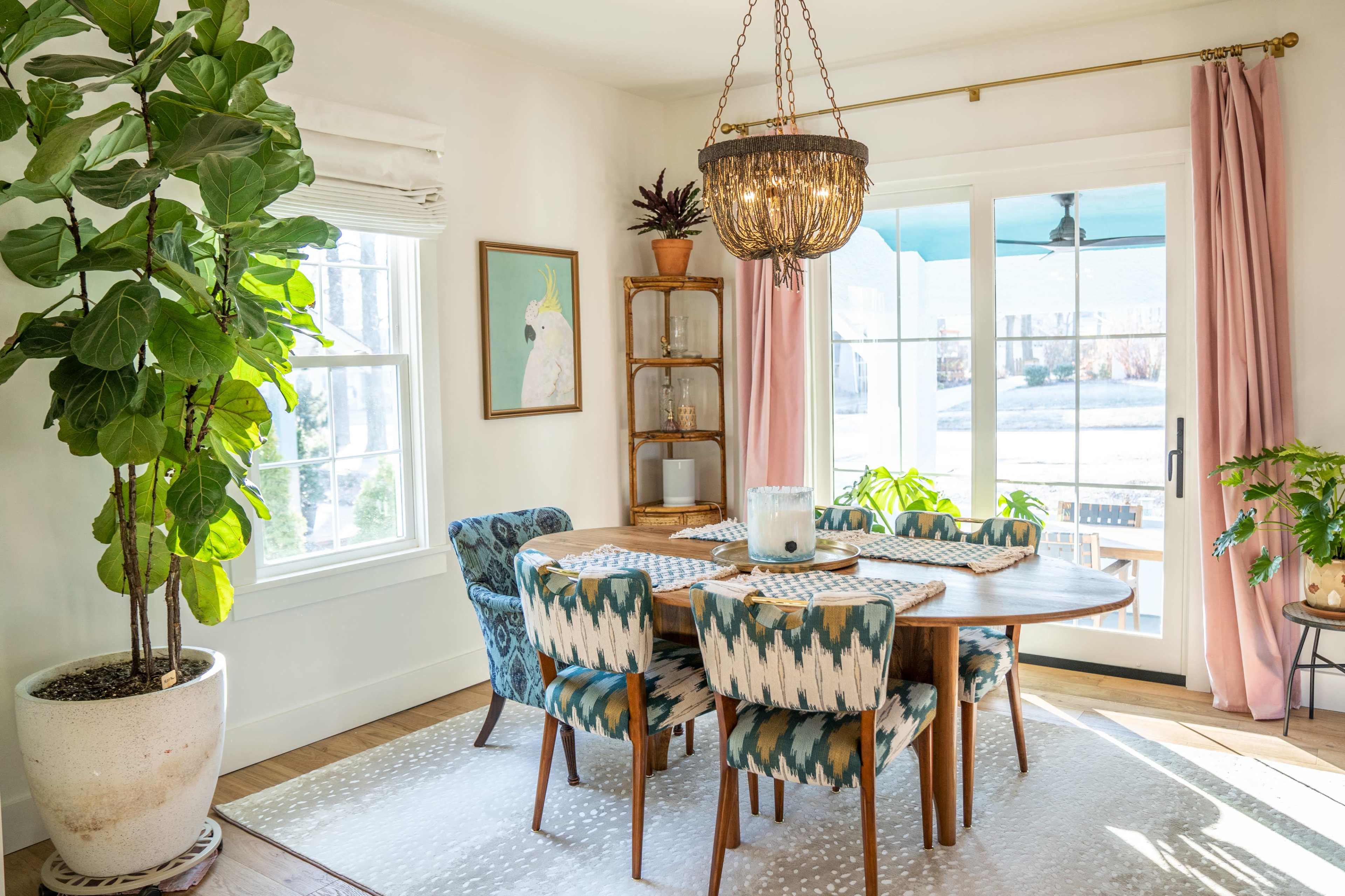 A dining area features a round table surrounded by patterned chairs, a chandelier above, a large potted plant, and sliding glass doors leading to an outdoor space.