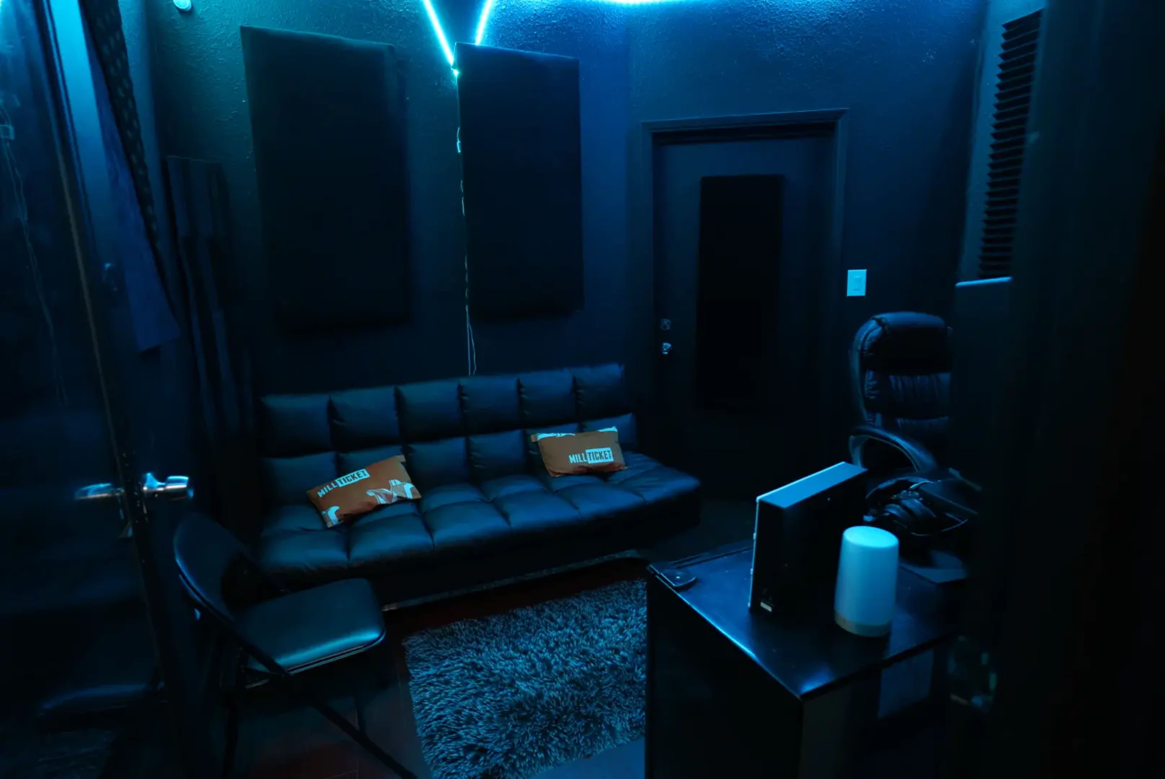 The image shows a small, dimly lit room with blue lighting, featuring a black sofa, a chair, and a desk with a lamp.