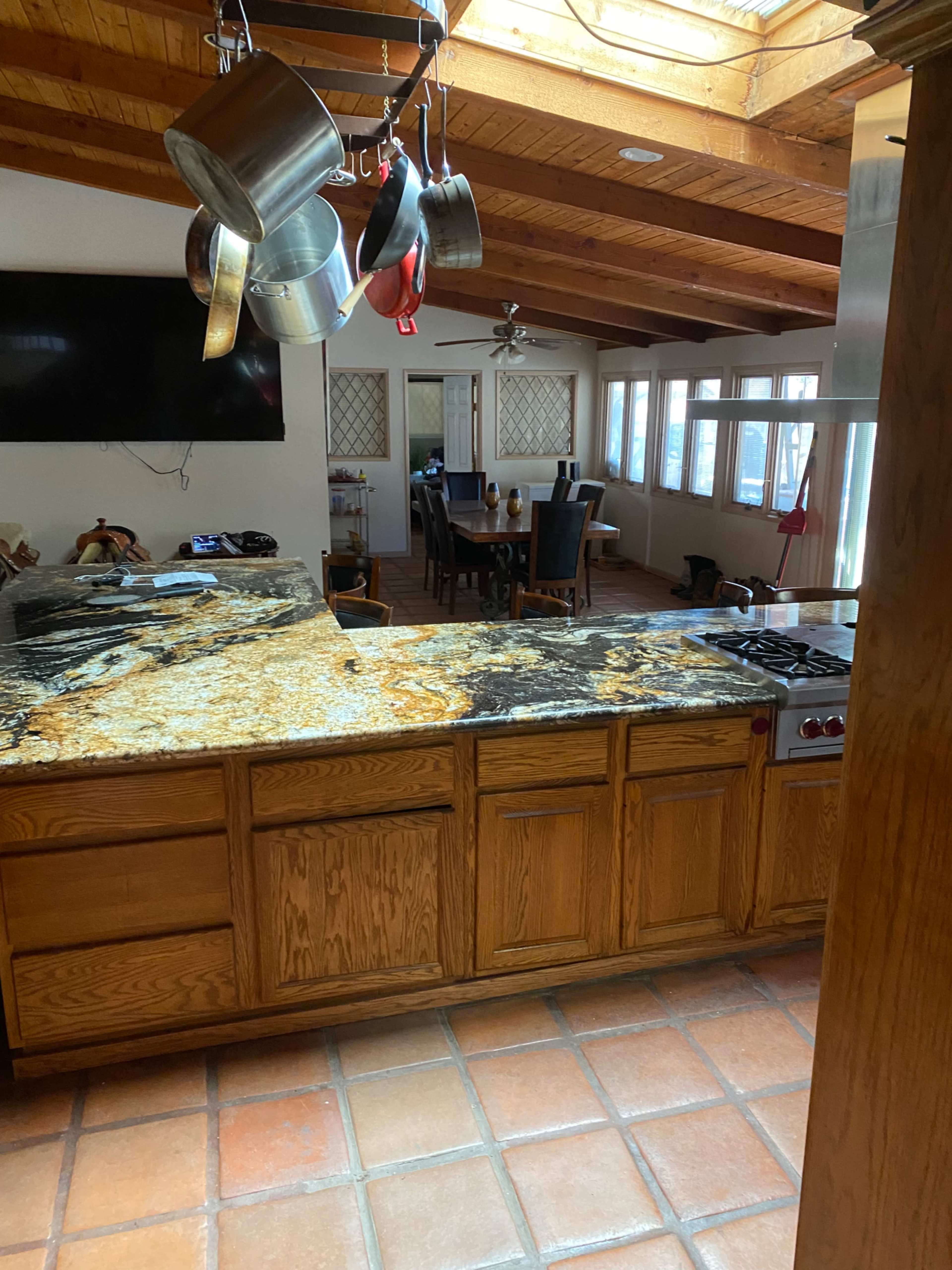 The image shows a kitchen with an expansive granite countertop, wooden cabinetry, and a view of a dining area through an open layout.