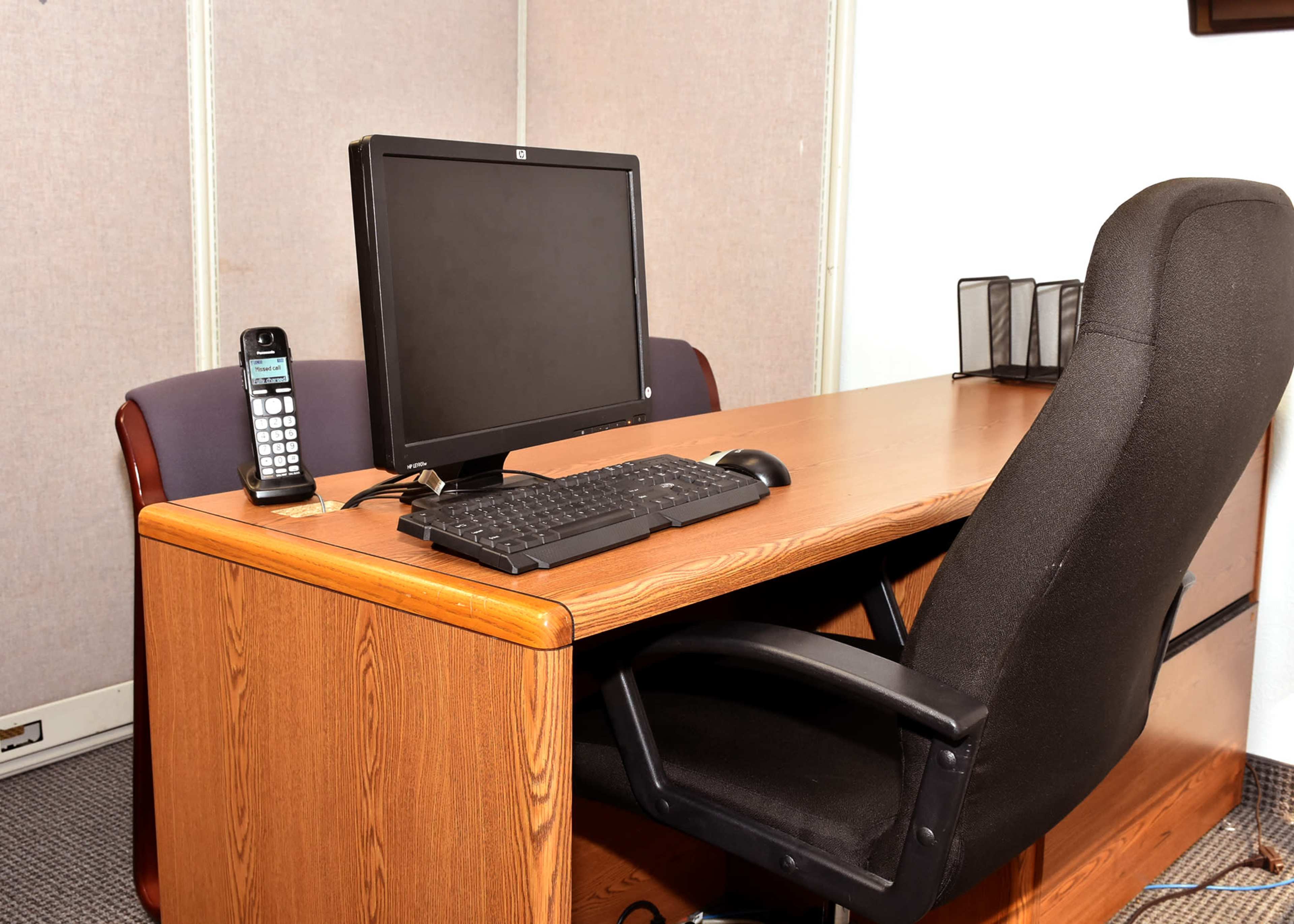 A wooden desk is set up with a computer monitor, a keyboard, a telephone, and an office chair in a cubicle.