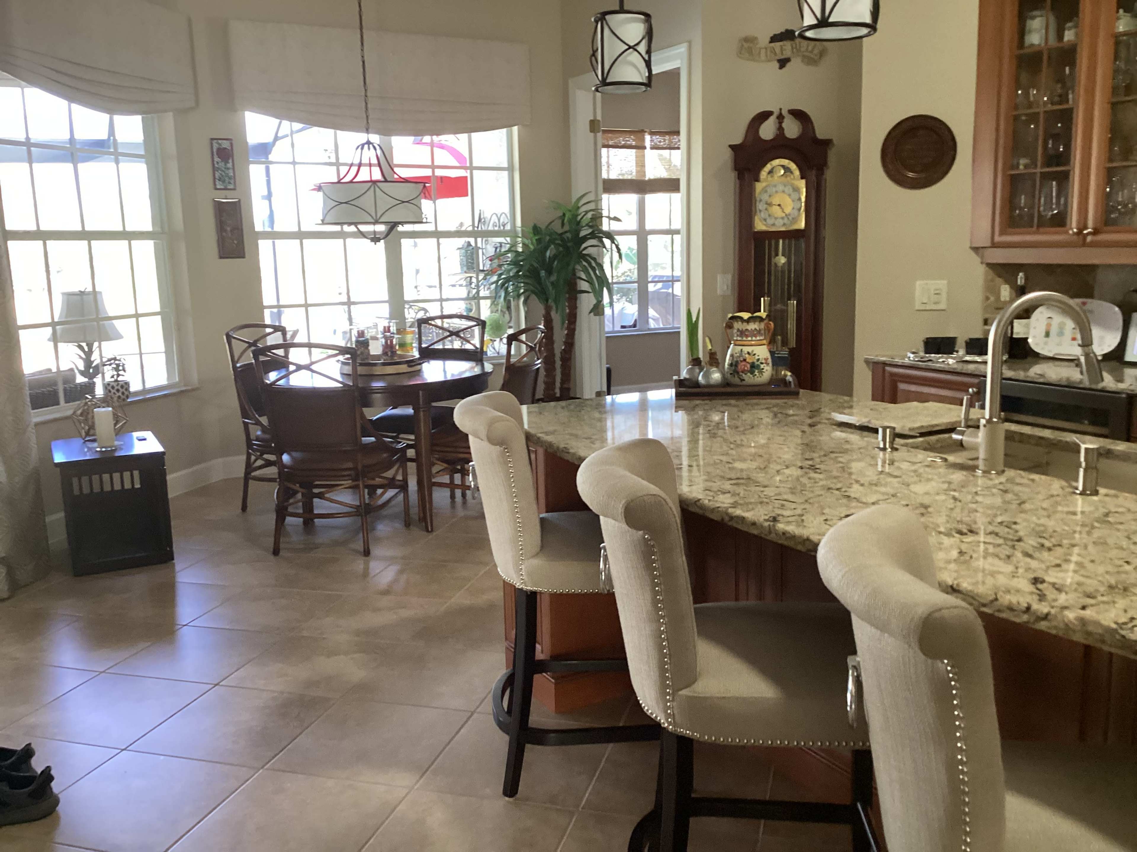 The image shows a kitchen area with a granite countertop, bar seating, and dining space filled with natural light from large windows.
