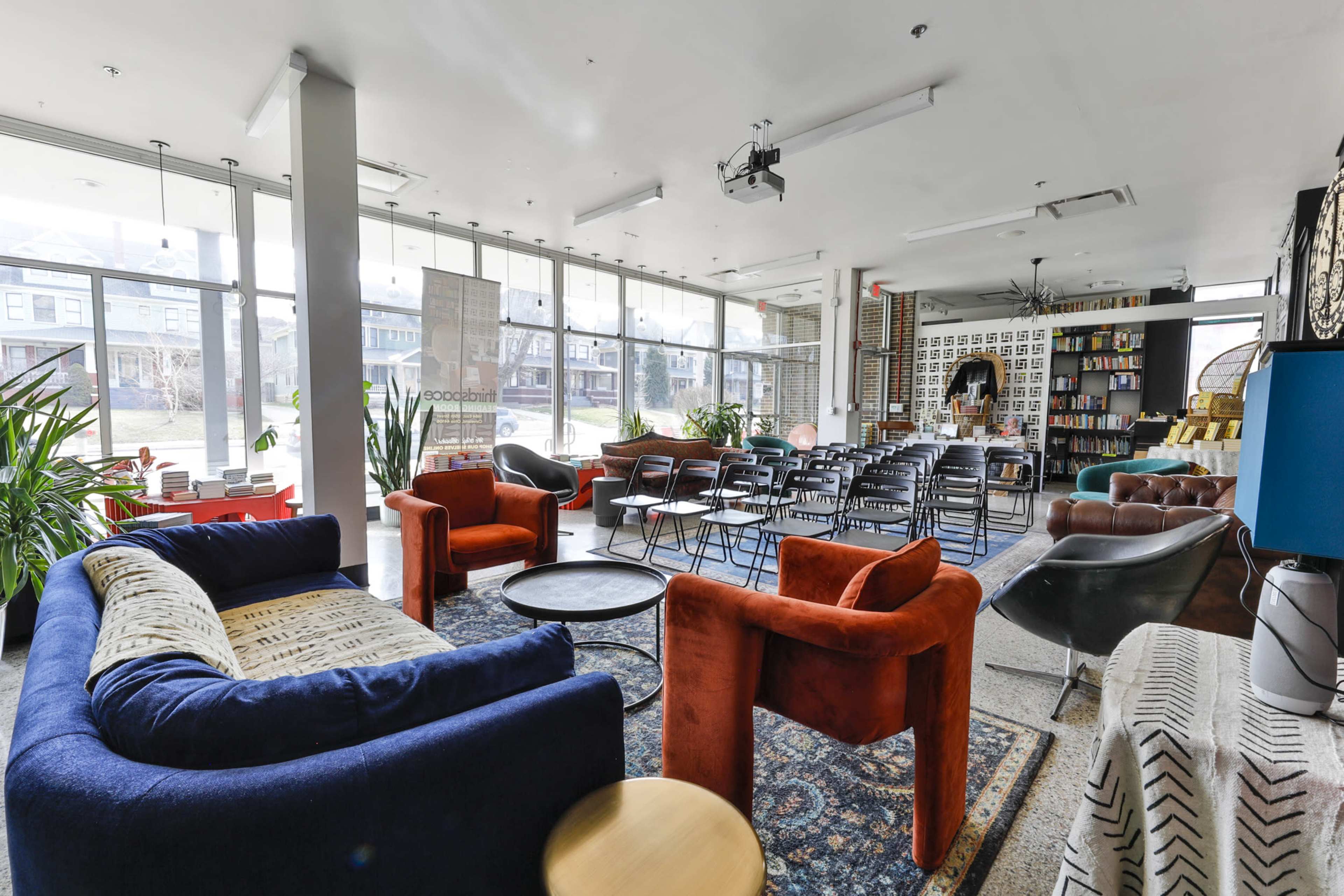 A brightly lit lounge area featuring various seating options, including blue and orange chairs, surrounded by large windows and a bookshelf.