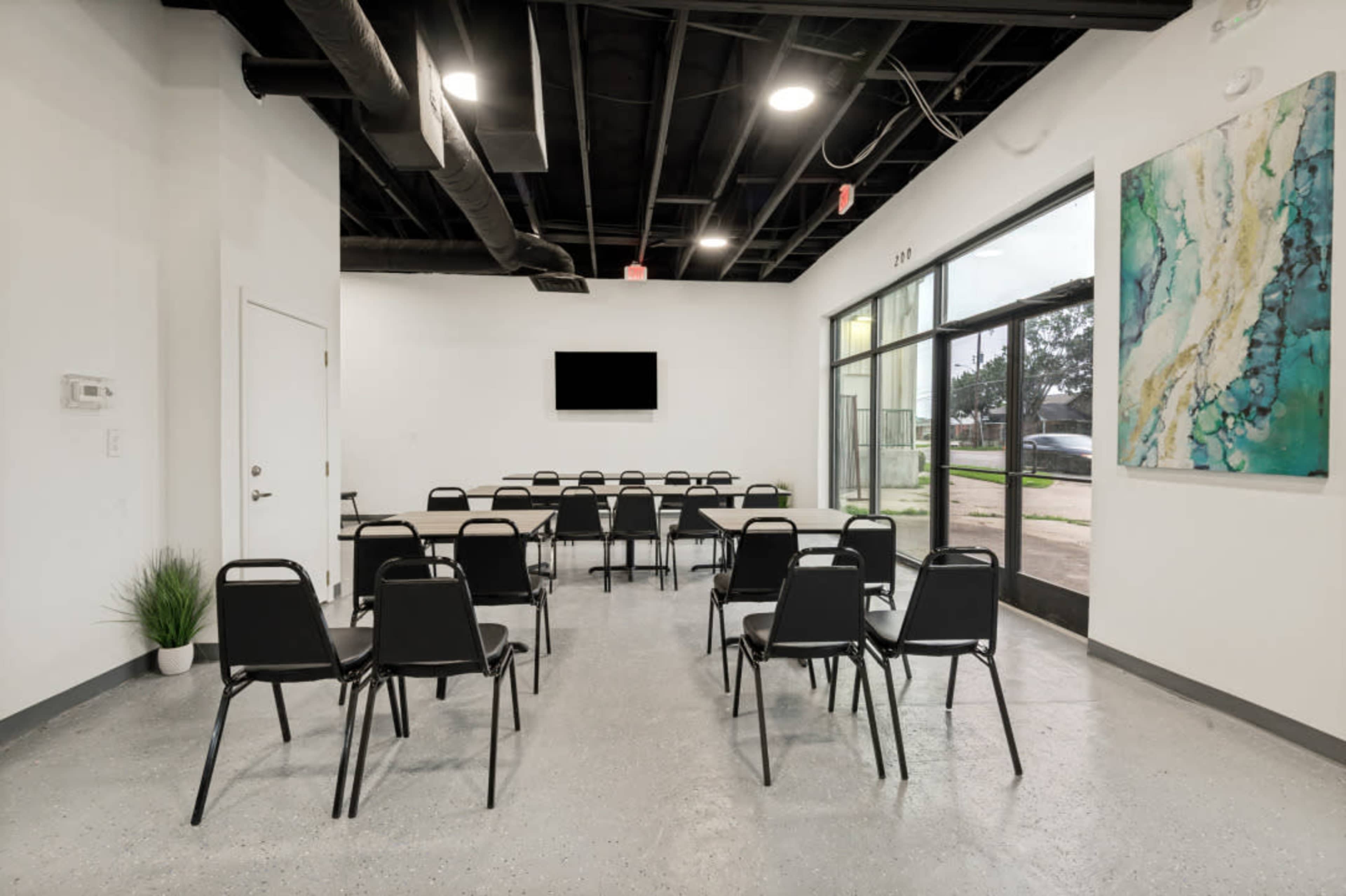 The image shows a spacious, brightly lit room arranged with several black chairs facing a wall-mounted screen.