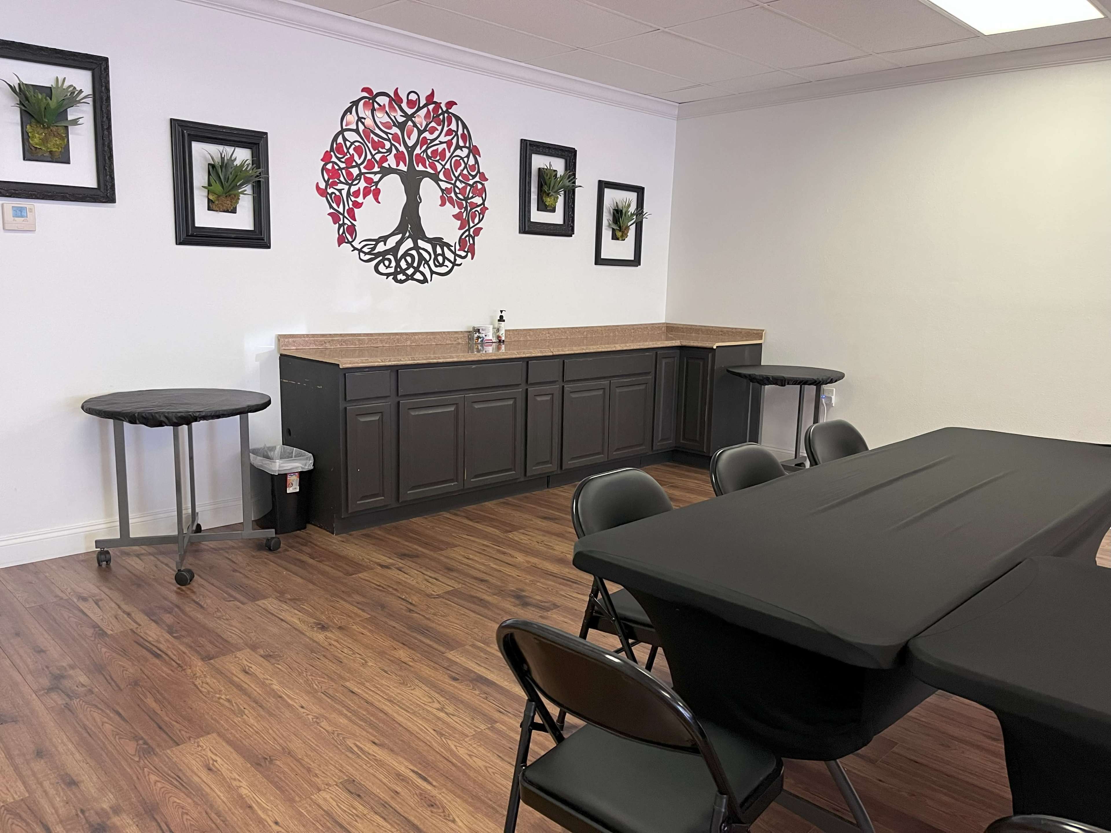 The room features a countertop with a black and red decorative wall piece, four black folding chairs, and two round tables against a white wall.