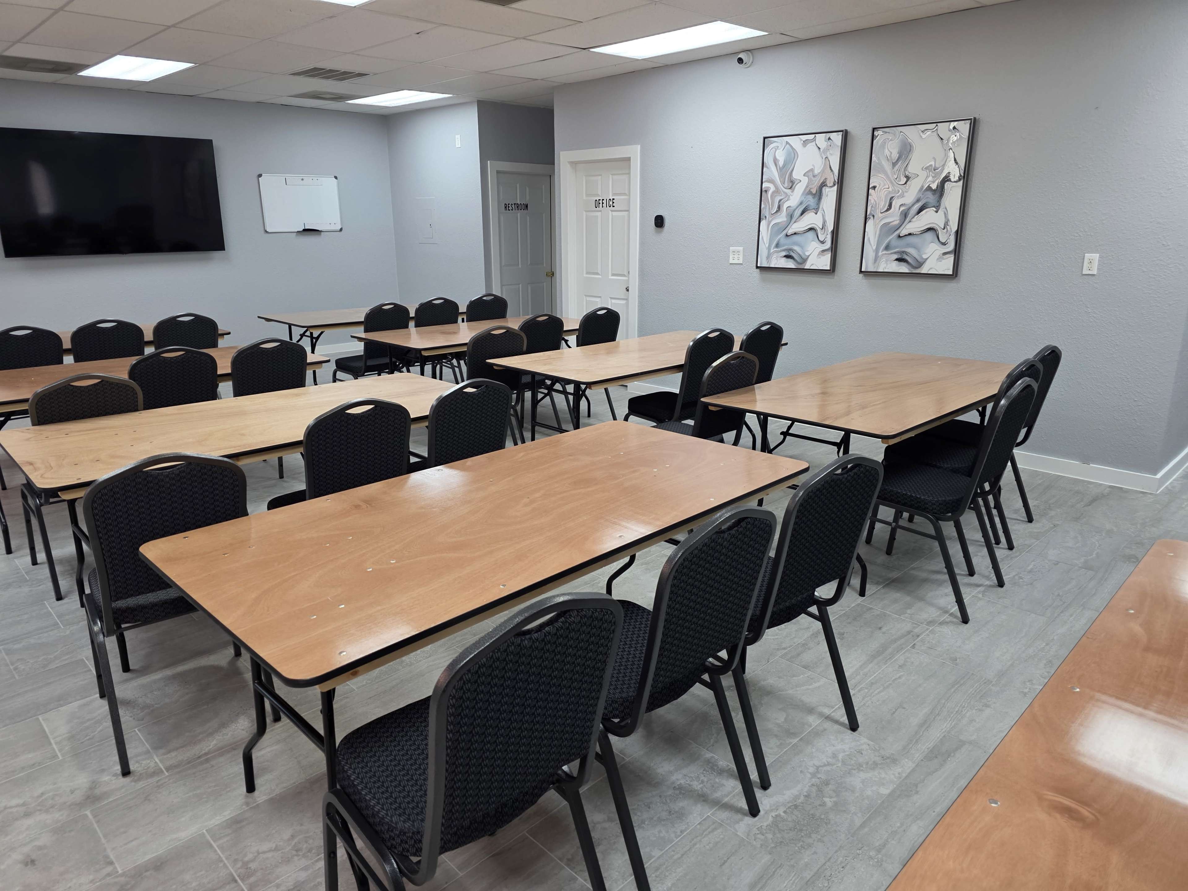 The image shows a meeting room with several wooden tables arranged in rows and black chairs placed around them.