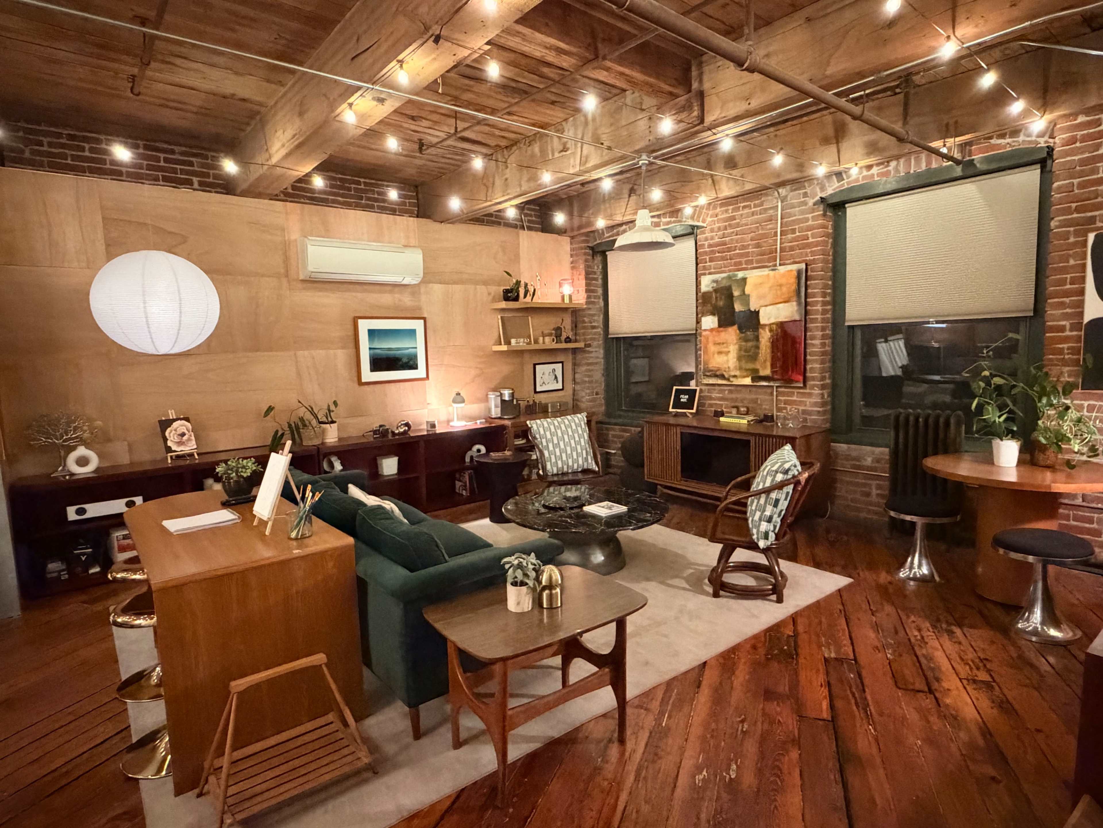 The image depicts a cozy office space featuring wooden beams, brick walls, various pieces of furniture, and warm lighting from overhead string lights.