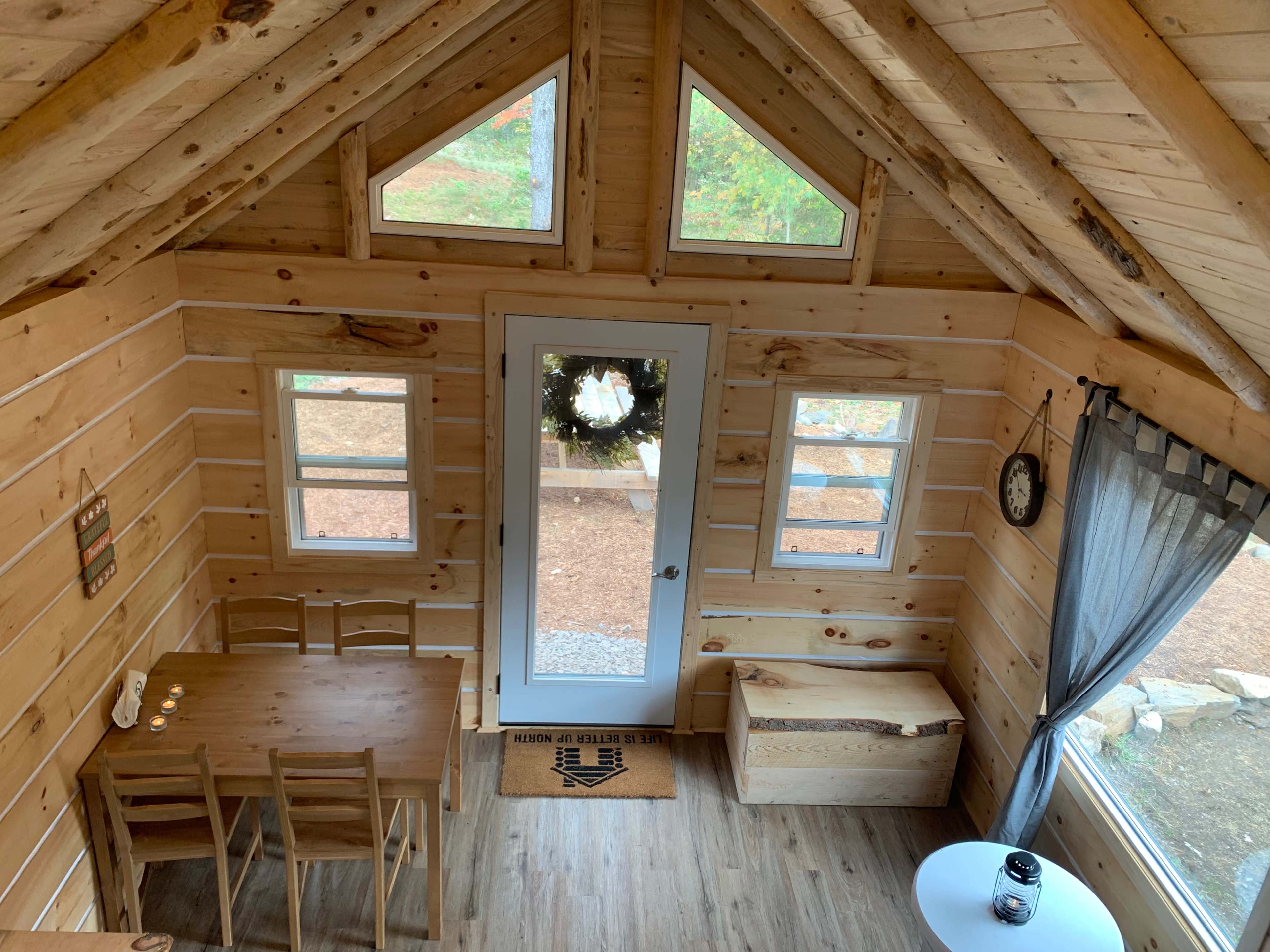 The image shows the interior of a wooden cabin with a triangular ceiling, featuring a dining table, chairs, and large windows providing natural light.