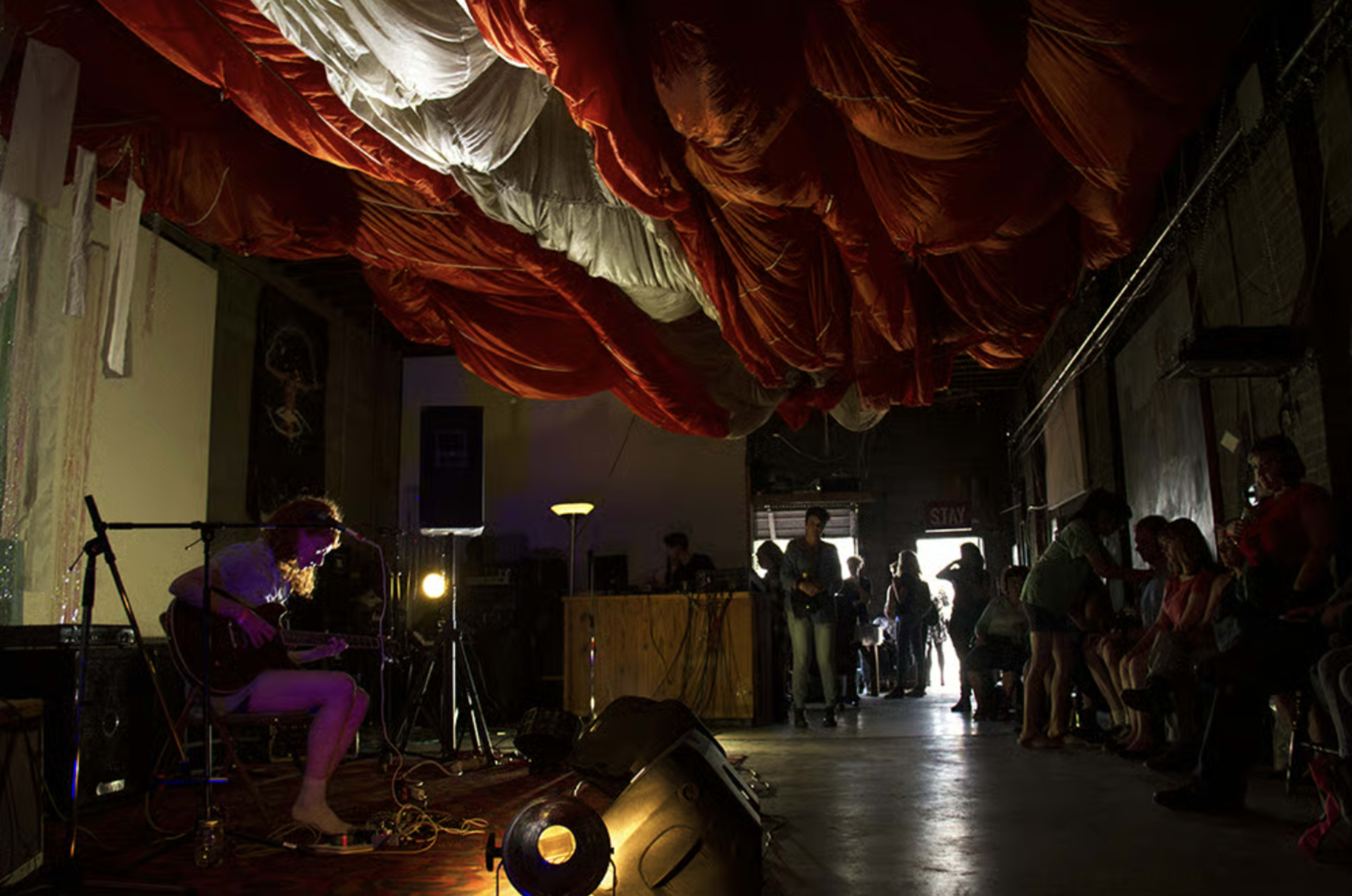 A musician plays guitar on stage in a dimly lit venue with a colorful fabric installation hanging from the ceiling and an audience seated nearby.