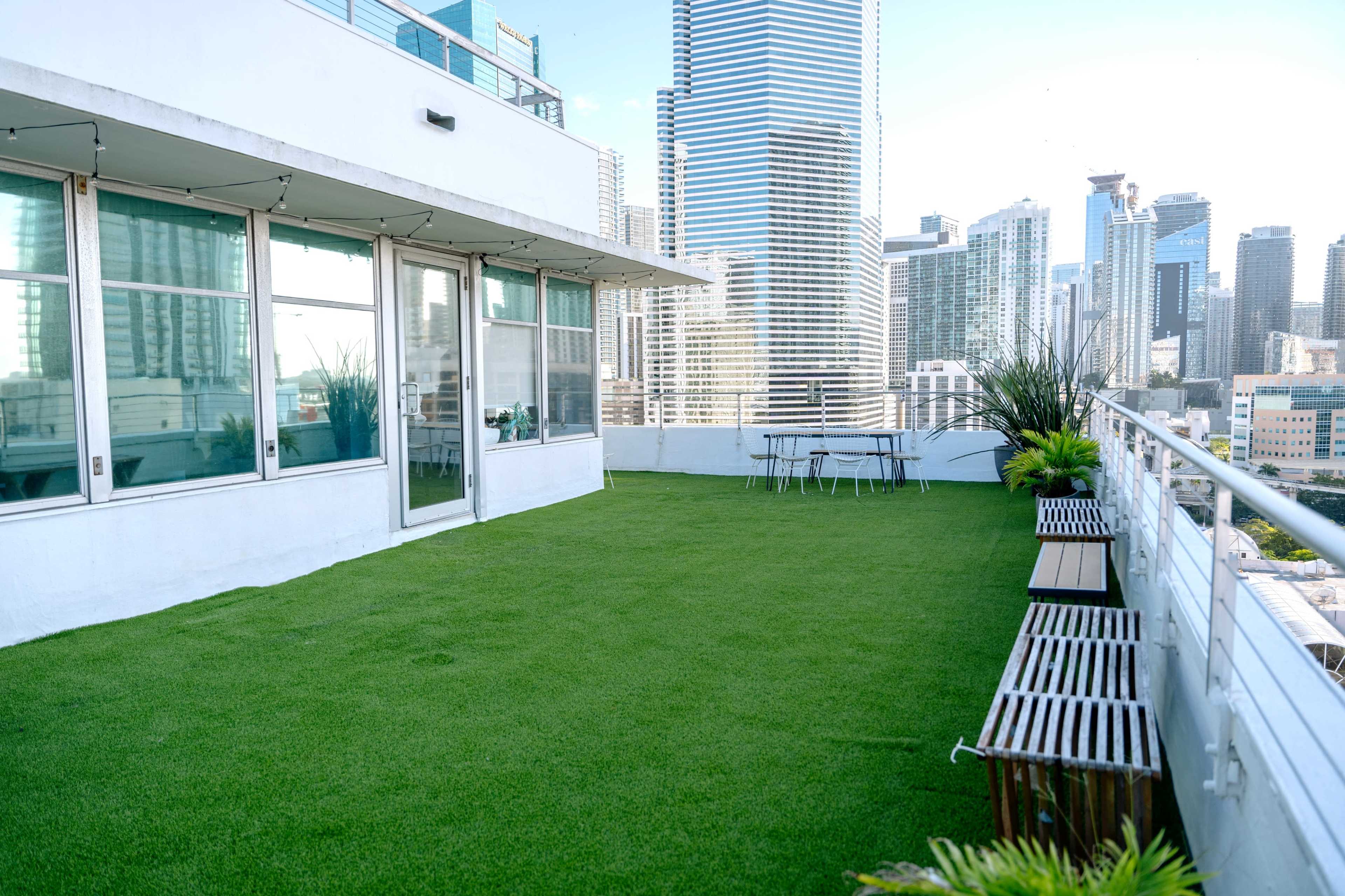 A spacious rooftop terrace features artificial grass, a table with chairs, and a view of tall city buildings.