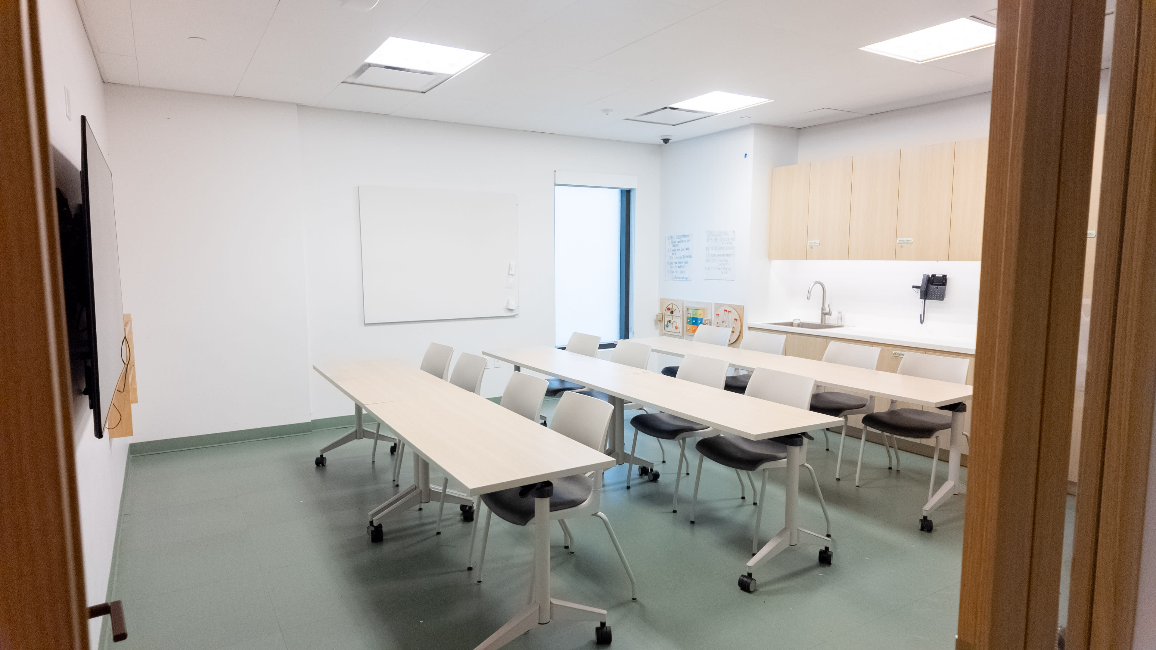 The image shows a modern classroom or meeting room equipped with a long table and several chairs, a whiteboard, and a kitchenette area.