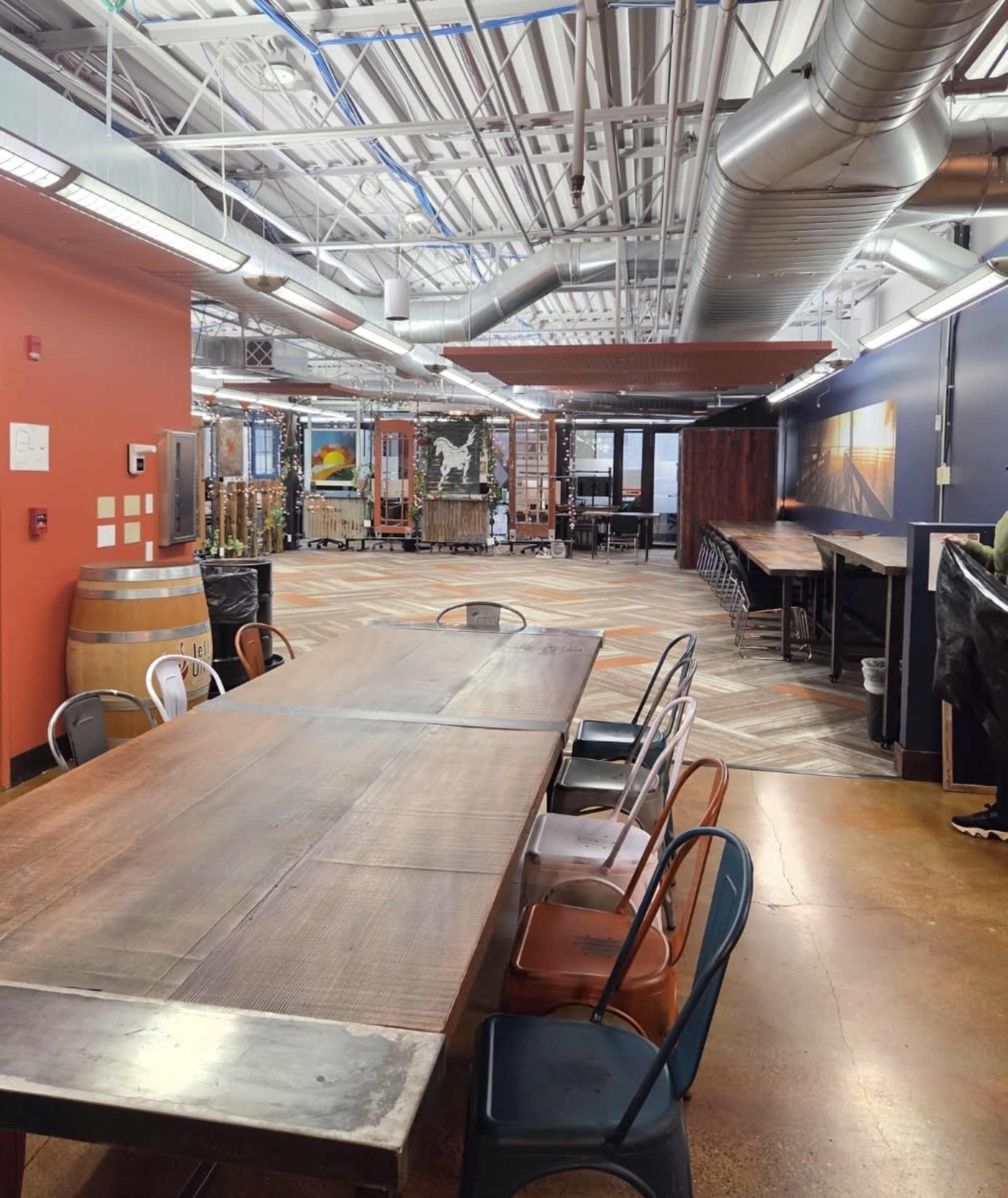The image shows a spacious dining area with a long wooden table surrounded by metal chairs, high ceilings, and industrial-style decor, including wooden barrels and a large mural.