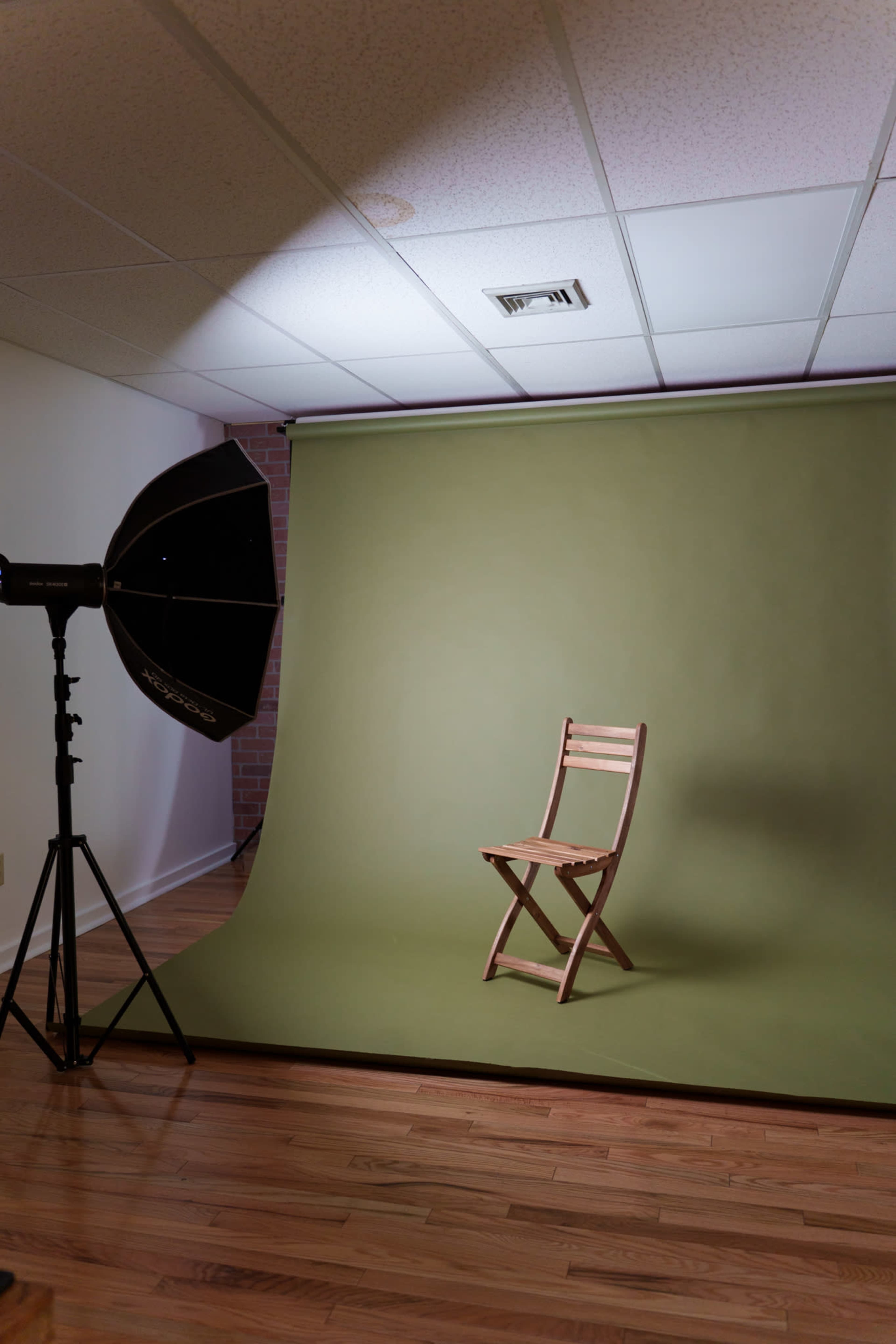 A wooden folding chair is positioned in front of a green backdrop, illuminated by a studio light on a stand.