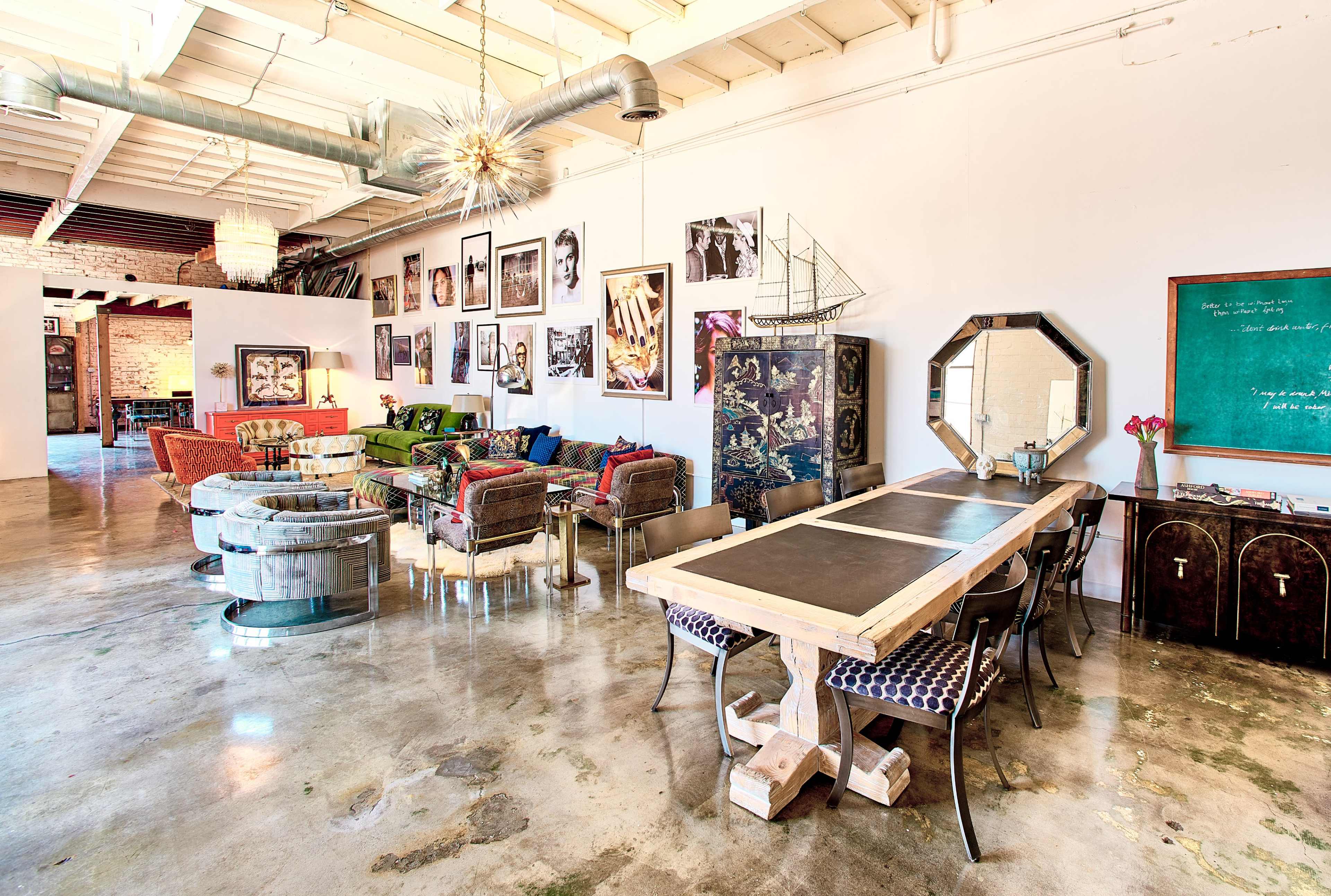 The image shows a spacious, modern interior with a wooden dining table, colorful seating areas, and framed artwork on the walls.