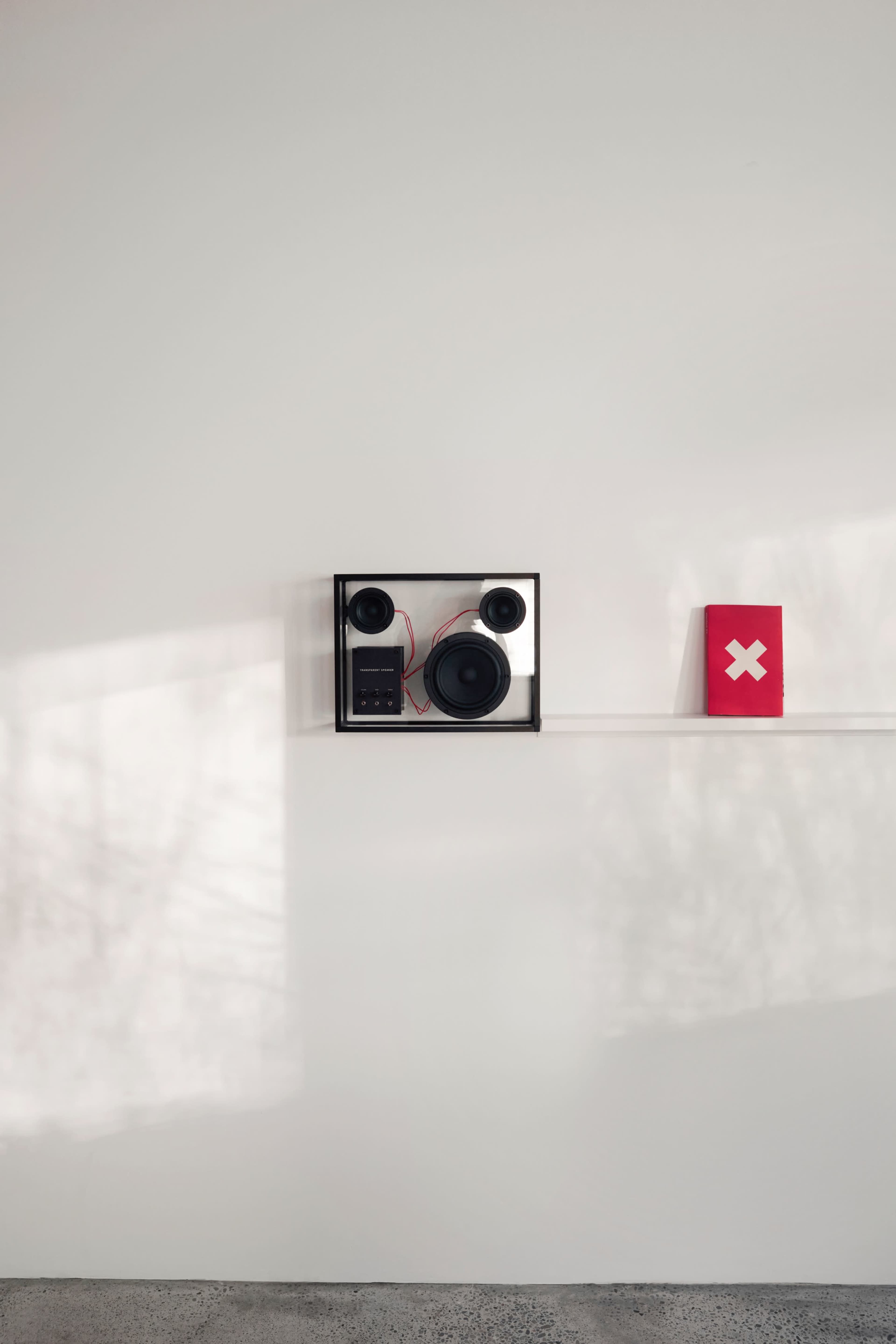 A stereo speaker system is mounted on a white wall beside a red book with a white cross symbol on its cover.
