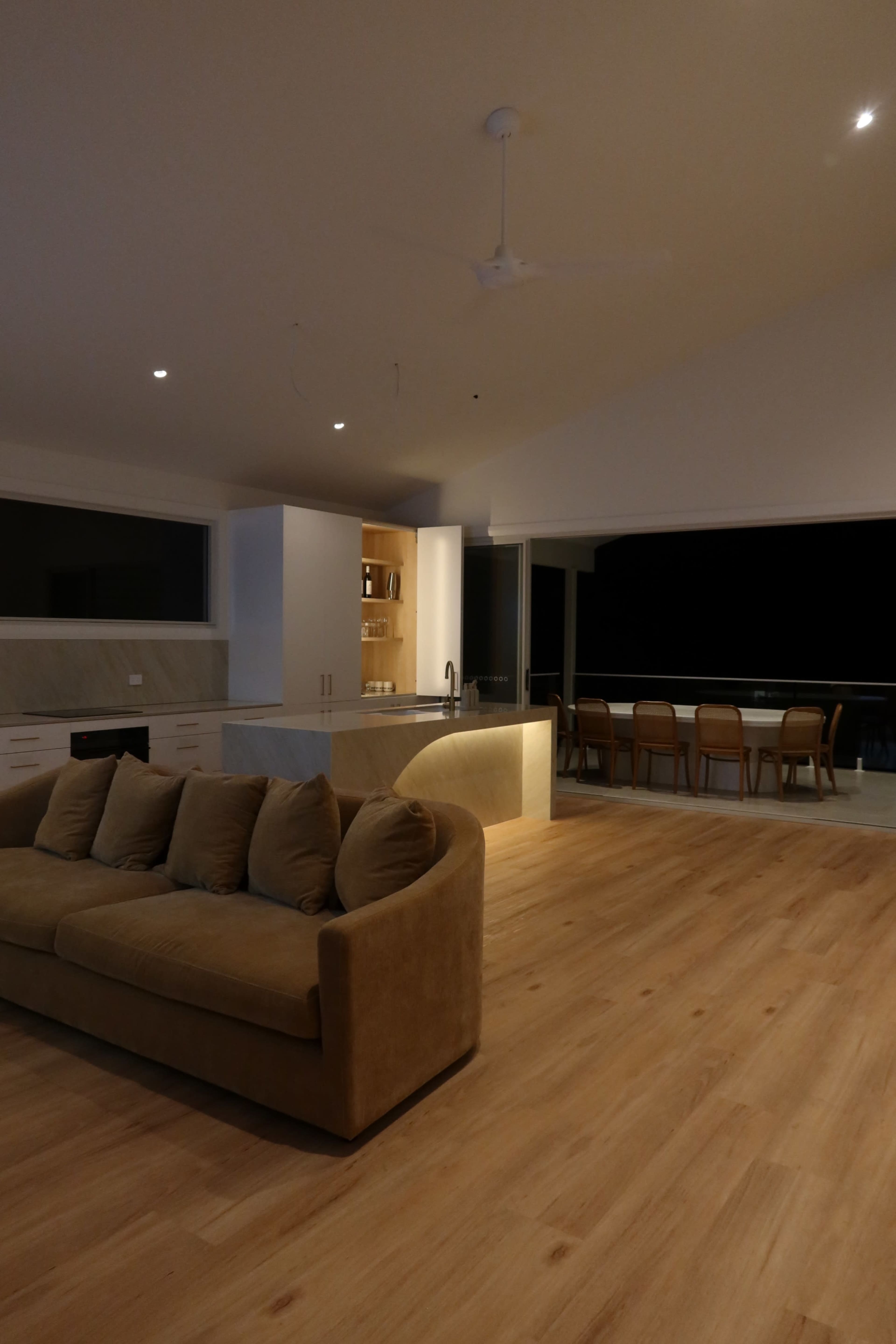 The image shows a modern, spacious living area featuring a light-colored sofa, a kitchen with a marble countertop, and a dining table with chairs visible in the background.